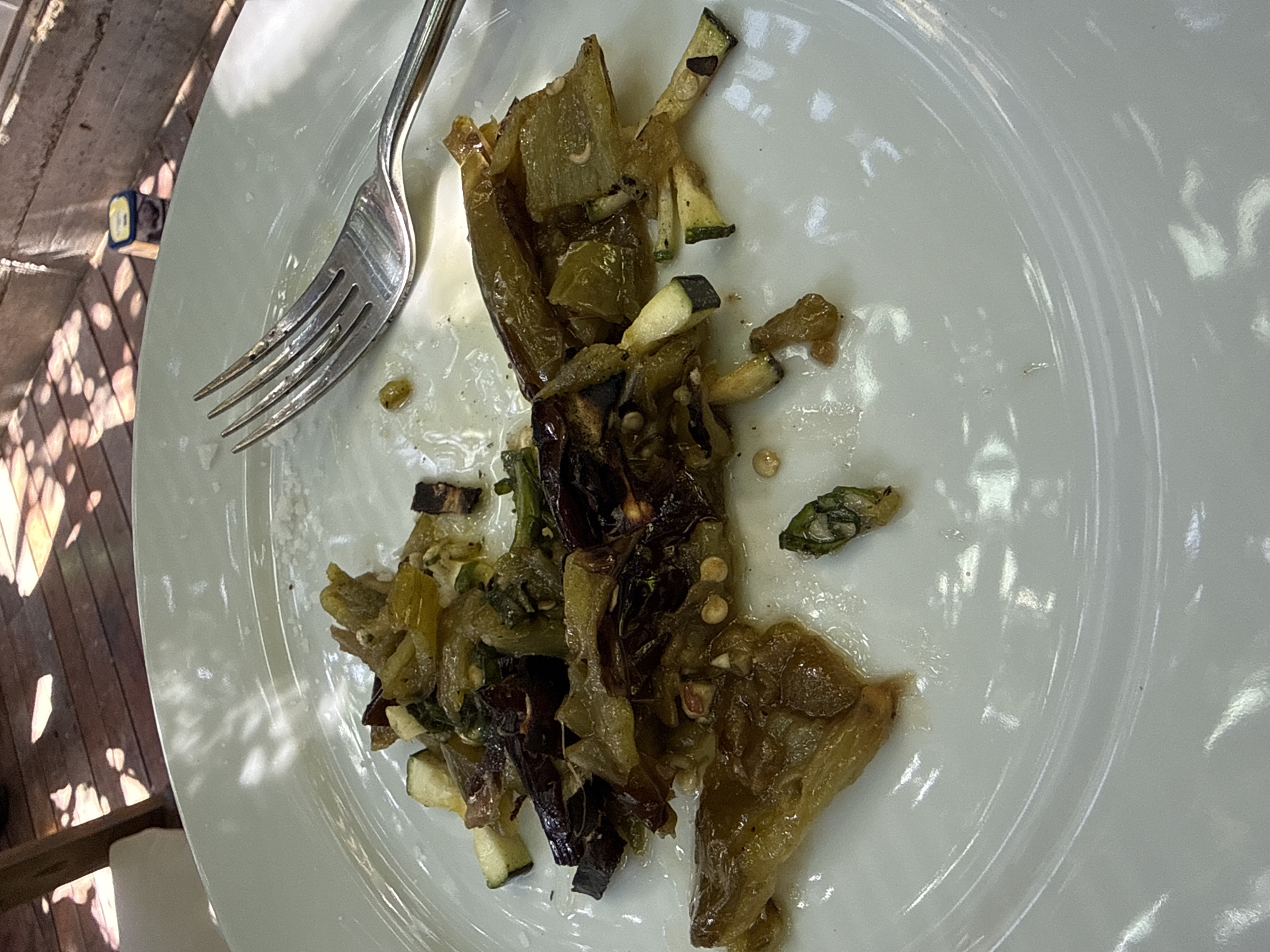 The image shows the remains of a meal on a white plate. The leftovers consist of cooked vegetables, including what appears to be peppers and zucchini. A fork rests on the plate next to the food. The scene is illuminated by dappled sunlight, suggesting it is outdoors, potentially on a patio or deck. The presence of wooden planks in the background further suggests an outdoor setting. The location in Barcelona, Spain is not evident in this image. There is no specific information about the presence of people, actions, or interactions.
