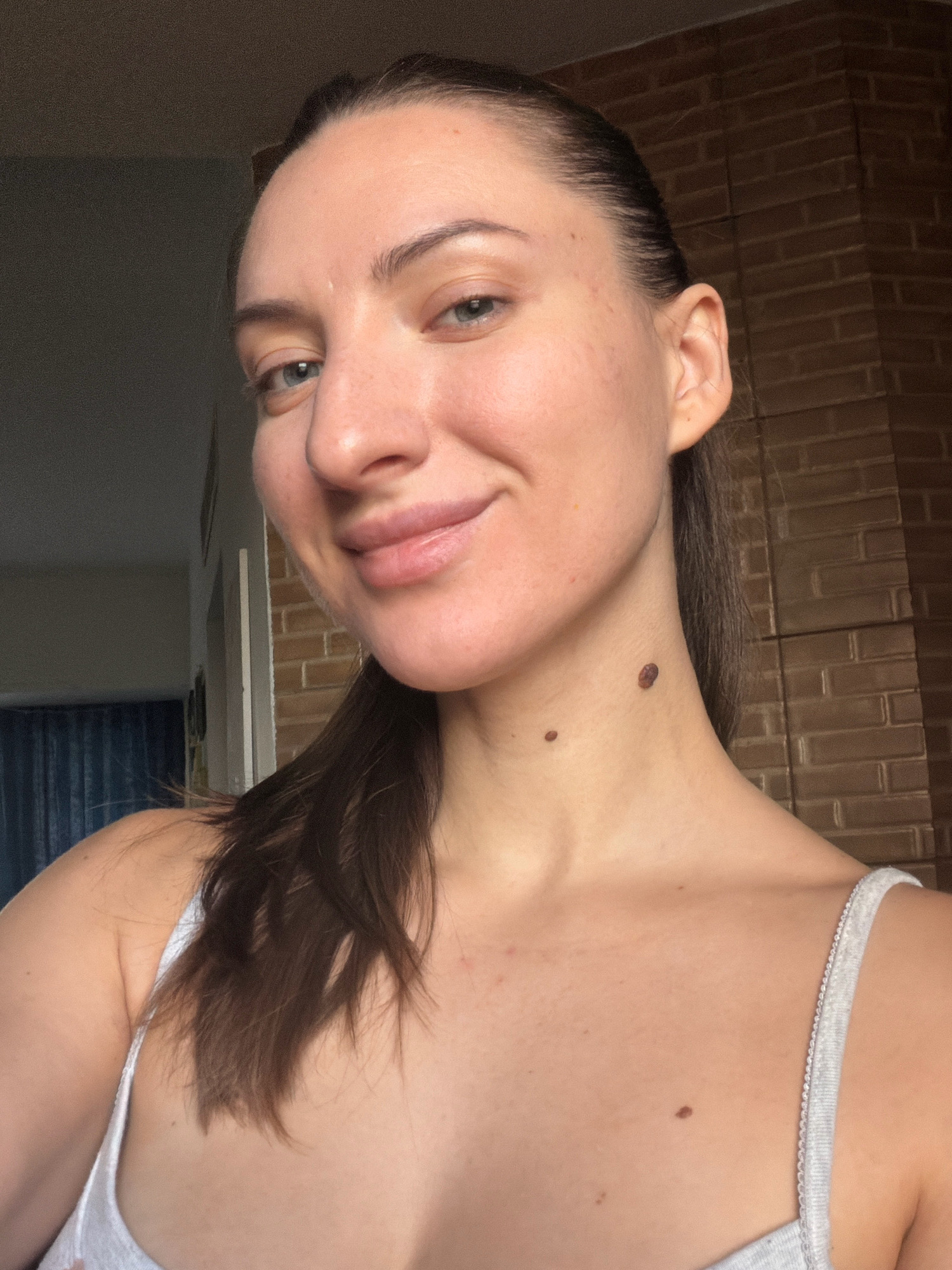 A young woman with brown hair pulled back in a ponytail smiles gently at the camera in a selfie. She is wearing a light-colored tank top. The background is a brick wall. The image is taken indoors.
