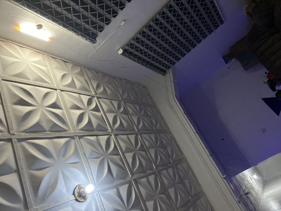 A close-up, low-angle view captures a textured white wall composed of square panels with floral patterns. A light fixture adorns the bottom of the wall. Above, the ceiling features a grid pattern. Part of the adjacent room shows a white wall illuminated by blue light. Furniture and toys are visible in the background.