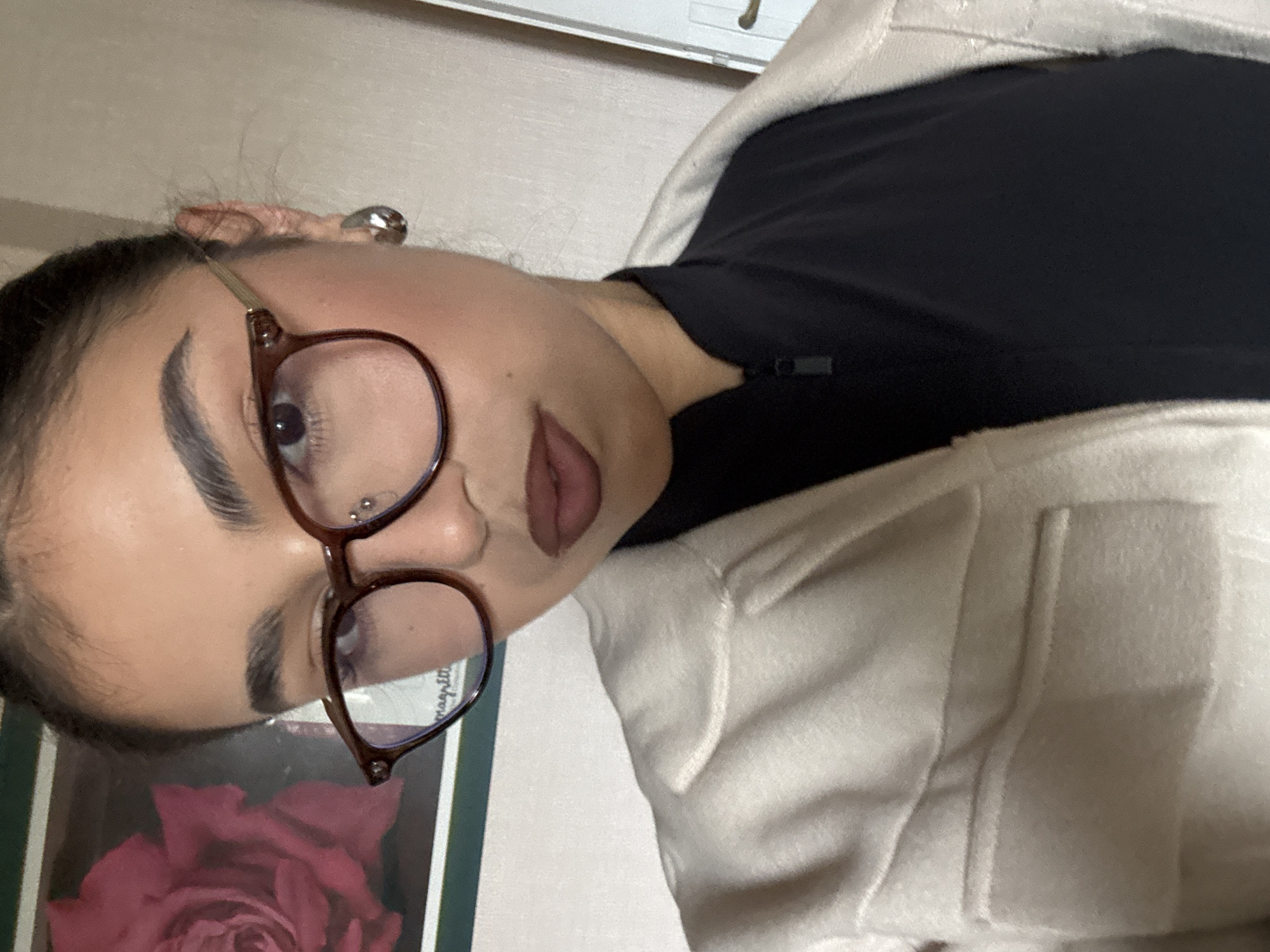 A person with fair skin, dark hair pulled back, and wearing glasses with round lenses is visible in a close-up, angled shot. The person is wearing a black top and a light-colored coat or jacket. There is a silver earring visible in their left ear, and a stud piercing on the left side of their nose. A picture with a red rose is partially visible near the bottom left corner of the frame. The background is indistinct but appears to be an interior setting.