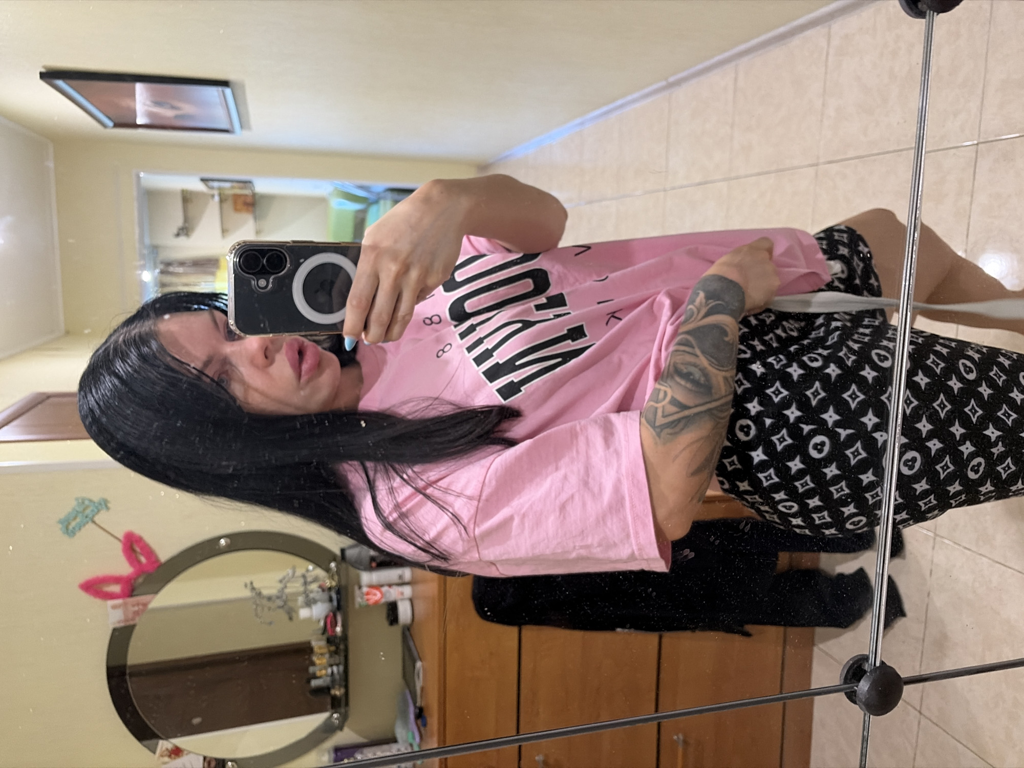 The media file shows a young woman with long black hair taking a mirror selfie. She is wearing a pink t-shirt with black lettering and black patterned shorts. The woman has tattoos on her left arm. The background appears to be a room with tiled floors and wooden furniture, possibly a bedroom or dressing area. A mirror is prominent in the frame, reflecting the woman and the surrounding environment. In the reflection, a framed picture is visible on the wall, and a decorative mirror is on a dresser. The lighting suggests it is daytime. There is no text clearly visible on the woman's clothing or in the immediate surroundings.