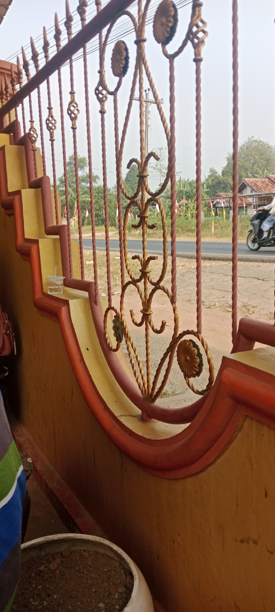 This is a photograph of a metal fence with ornate scrollwork, possibly in Gondang Rejo, Indonesia, as suggested by the Indonesian flags visible in the background. The fence is painted a reddish-brown color, with gold accents on the decorative elements. Behind the fence, a road is visible with a motorcycle in motion. The environment appears to be somewhat rural, with trees and houses lining the road. In the foreground, part of a yellow and red painted structure, possibly a staircase or wall, is visible. A clear plastic cup with a straw and a small amount of liquid sits on one of the yellow steps. The sky is overcast.