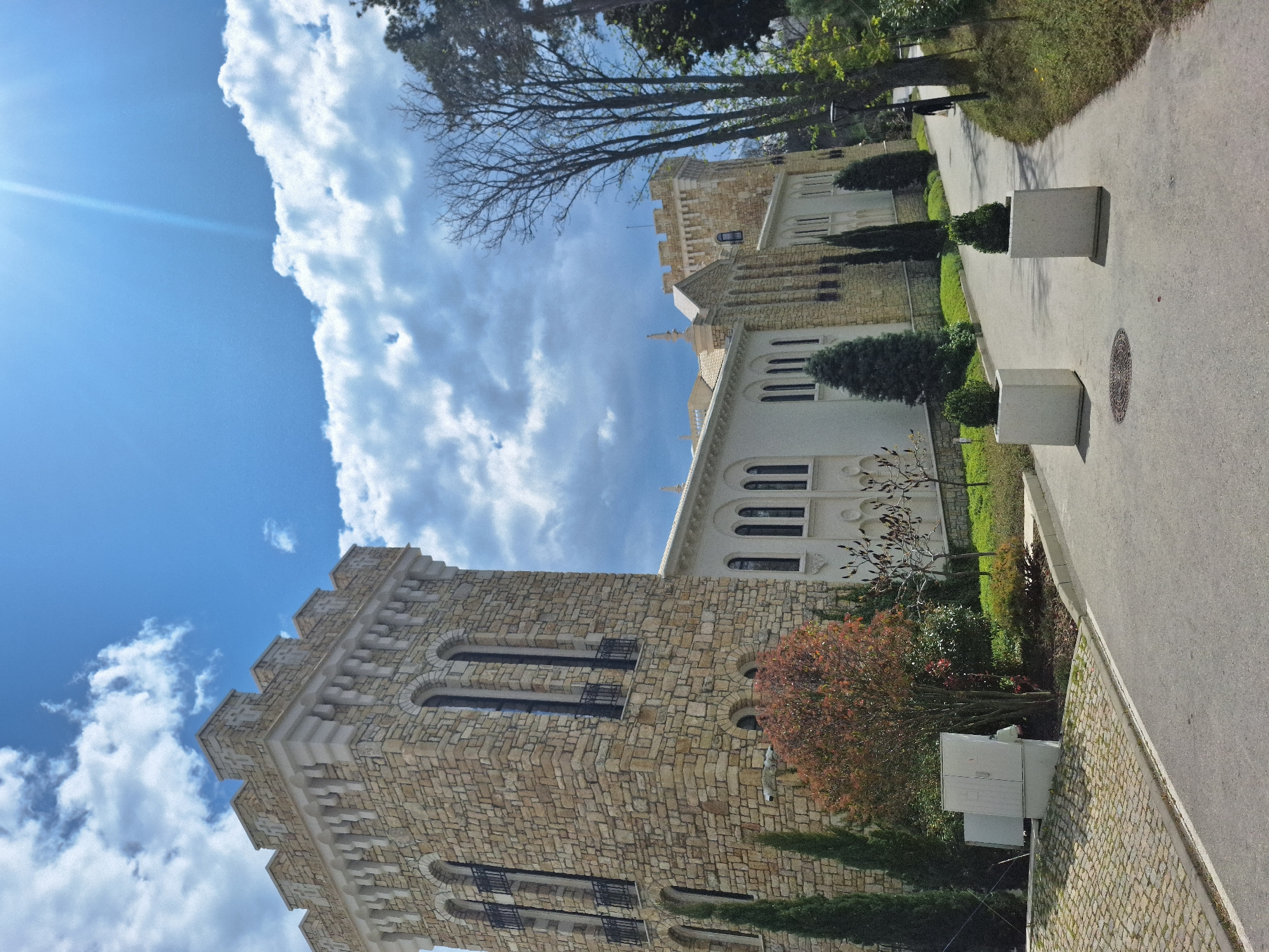 The image captures a stone building under a blue sky with scattered clouds. The building has multiple arched windows and is likely a historic structure, featuring a tower-like extension. The architecture suggests a possible castle or governmental building.
Located in Varna, Bulgaria, a pathway runs alongside the building, bordered by vegetation including trees, shrubs, and grass. Cement planters are placed along the path. The environment implies an urban setting with a blend of nature and architecture.