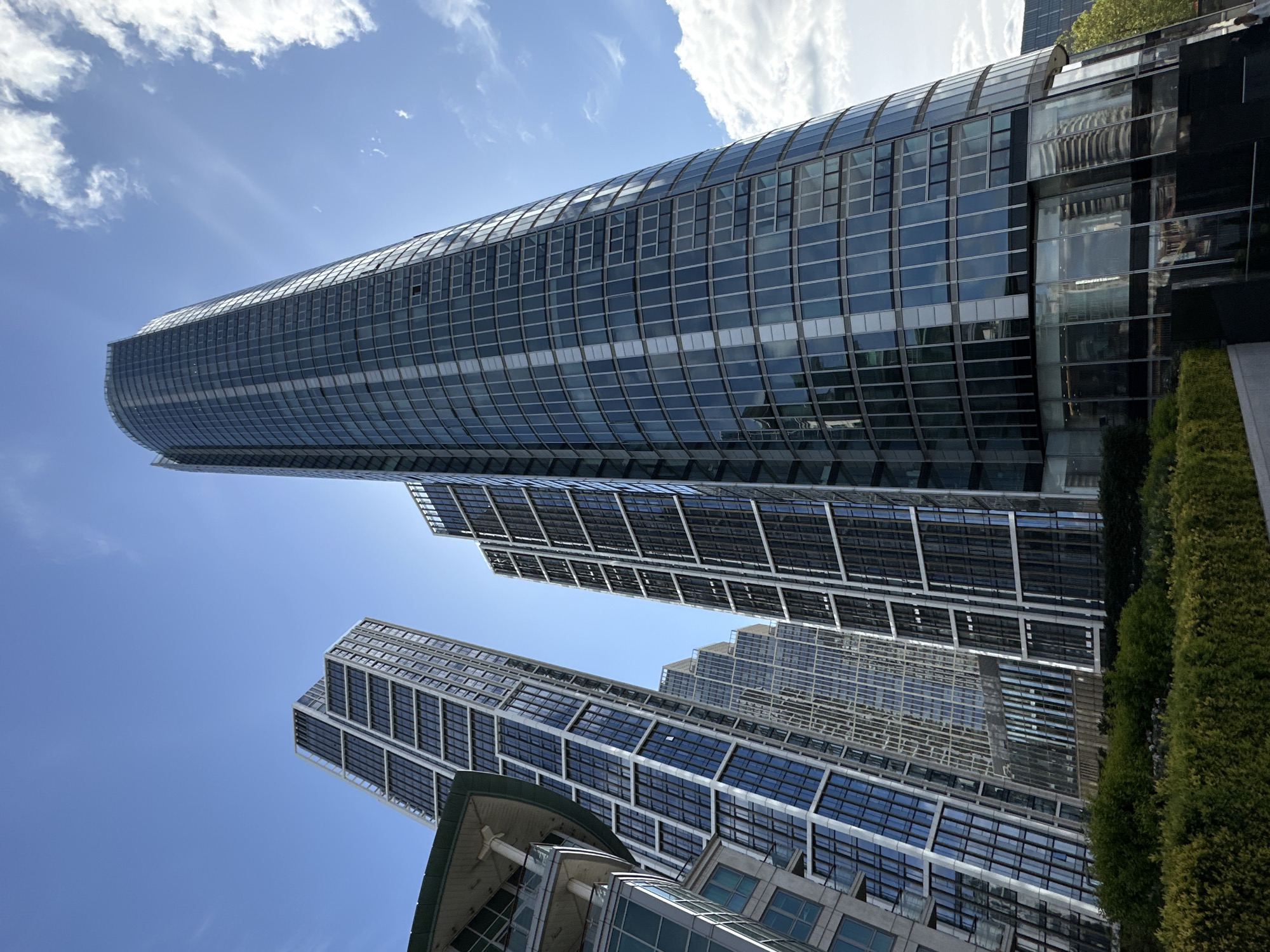 The image shows a low-angle view of modern skyscrapers in London, United Kingdom. The buildings are characterized by glass facades and linear architectural designs. The sky is blue with scattered white clouds. Green shrubbery is visible on the right side of the frame, suggesting a garden or landscape feature at the base of the buildings.