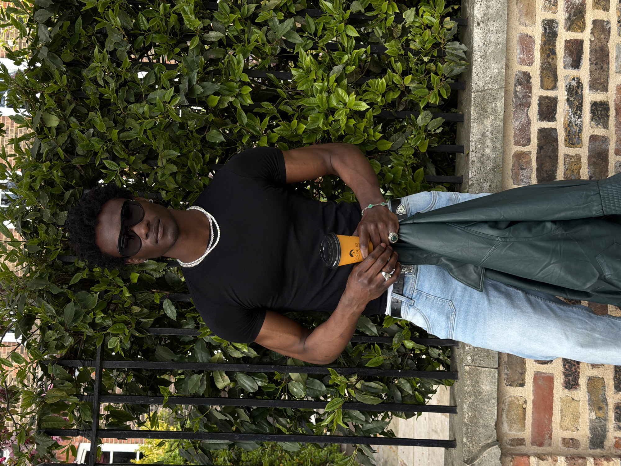 The photograph depicts a stylishly dressed individual, likely male, outdoors, possibly in London, United Kingdom. The subject is wearing a black t-shirt, light blue jeans, a silver chain, sunglasses, and rings, holding a takeaway coffee cup in one hand and a green leather jacket in the other. He stands in front of a wrought iron fence with dense green foliage behind it. Adjacent to the fence is a brick wall. The photograph is taken from a slightly elevated angle and appears to be in a residential or commercial area.