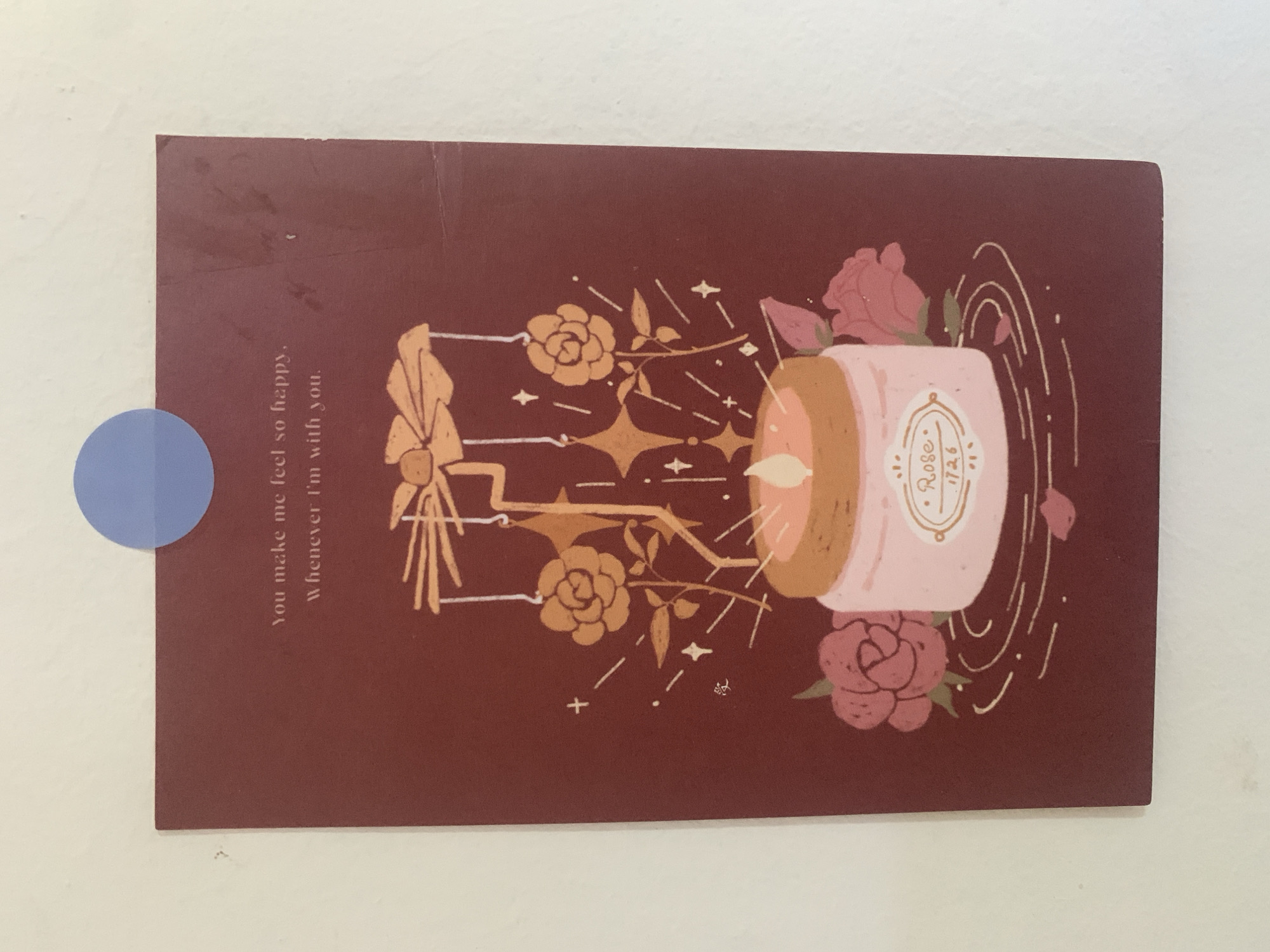 A postcard with a burgundy background is affixed to a white wall with a blue circular sticker. The postcard features an illustration of a lit candle in a pink jar, surrounded by pink roses. Above the candle, there is a mobile with star and flower shapes. Text on the postcard reads: "You make me feel so happy. Whenever I'm with you." There is also a small Asian character on the left side of the postcard. The postcard is slightly bent at the top right corner. The image does not contain any identifiable landmarks of Kuala Pembuang, Indonesia.