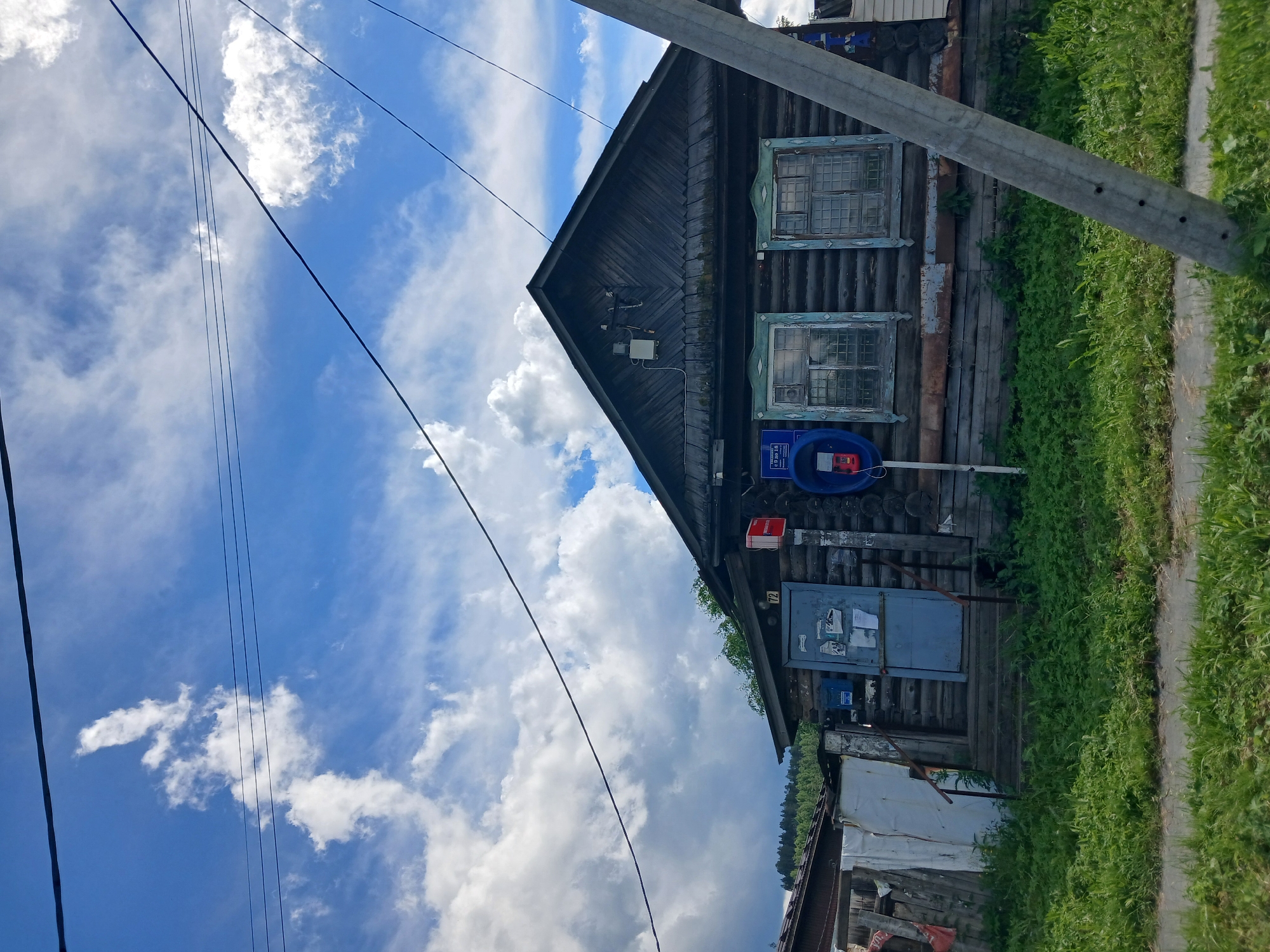 The image depicts a wooden log house in the village of Staroye Reshyoty, Russia. The house has a dark wooden facade with two windows, each with blue decorative trim. A blue door is located on the left side of the house, leading to a set of wooden stairs. Next to the door, there is a mailbox and a payphone booth with a red phone inside. Above the door, a sign with the number "72" is visible. Overhead, power lines stretch across a blue sky with scattered white clouds. The foreground is filled with green grass and a paved pathway. To the left of the main house, a smaller structure with a white tarp is visible. The overall impression is that of a rural, possibly aging, residential building.