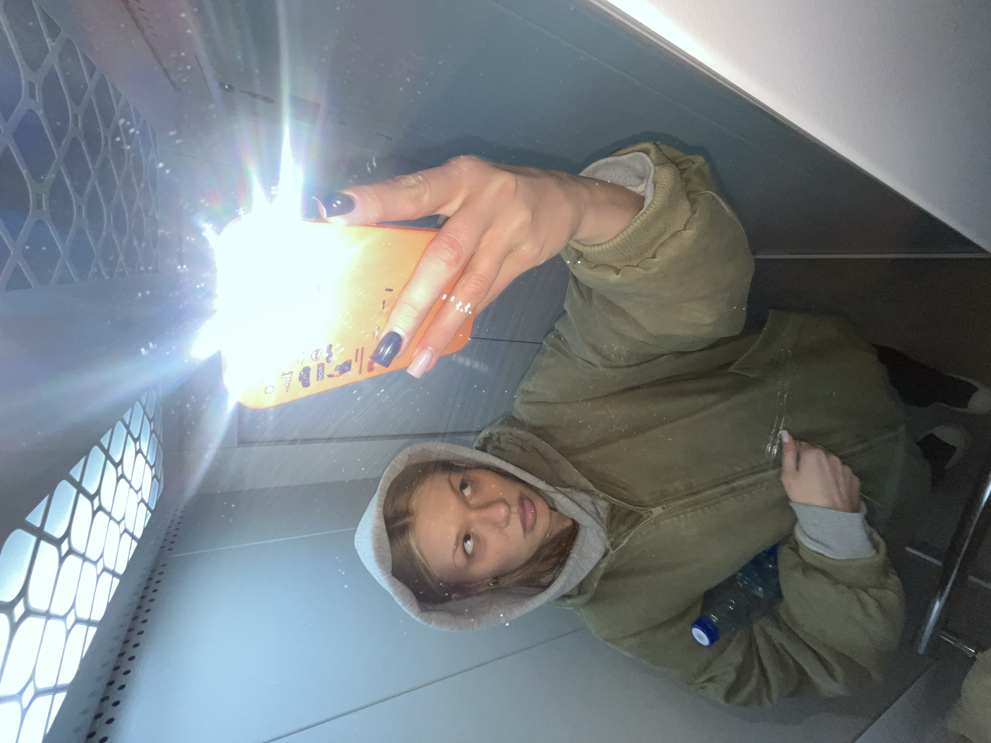 A woman is holding an orange container up to the light near a barred window in what appears to be a small room or enclosure. She wears a hooded sweatshirt beneath a green jacket. The background is indistinct, showing sections of wall and floor. A water bottle is partially visible near her waist. The location is unknown.
