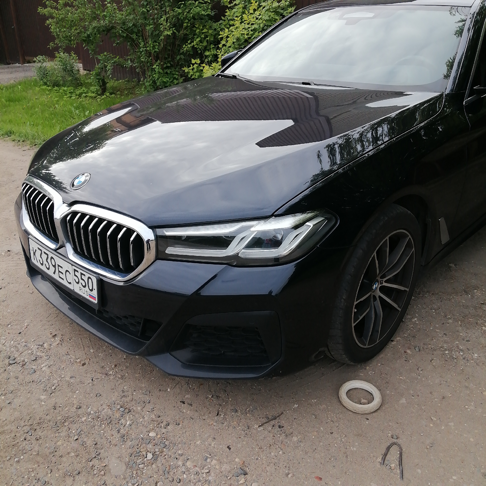 A black BMW 5 Series sedan (G30 generation) is prominently featured, parked on a dirt and gravel surface. The vehicle's front and right side are visible, angled towards the left. The car's highly reflective black paint shows reflections of green foliage, a dark fence or wall, and the sky. It sports a distinctive chrome kidney grille and angular LED headlights, with the text "VR LED" partially visible on the right headlight.
The license plate, mounted on the front bumper, reads "K339EC 550. RUS", indicating its registration in Russia, specifically Omsk Oblast (region 550). The car is equipped with multi-spoke alloy wheels.
The setting is outdoors during the daytime under clear or partly cloudy skies, as evidenced by the bright lighting and reflections. The background consists of lush green bushes and trees, along with a dark, corrugated structure, possibly a fence or building. The ground directly beneath and around the car is unpaved, composed of light brown dirt and various sizes of gravel and small stones.
On the ground near the front passenger-side wheel, there's a white, tire-shaped object, possibly a toy, and a U-shaped piece of metal sticking out of the ground. There are no people present, and the activity is limited to the car being stationary. The environment suggests a casual, possibly rural or suburban outdoor parking spot.