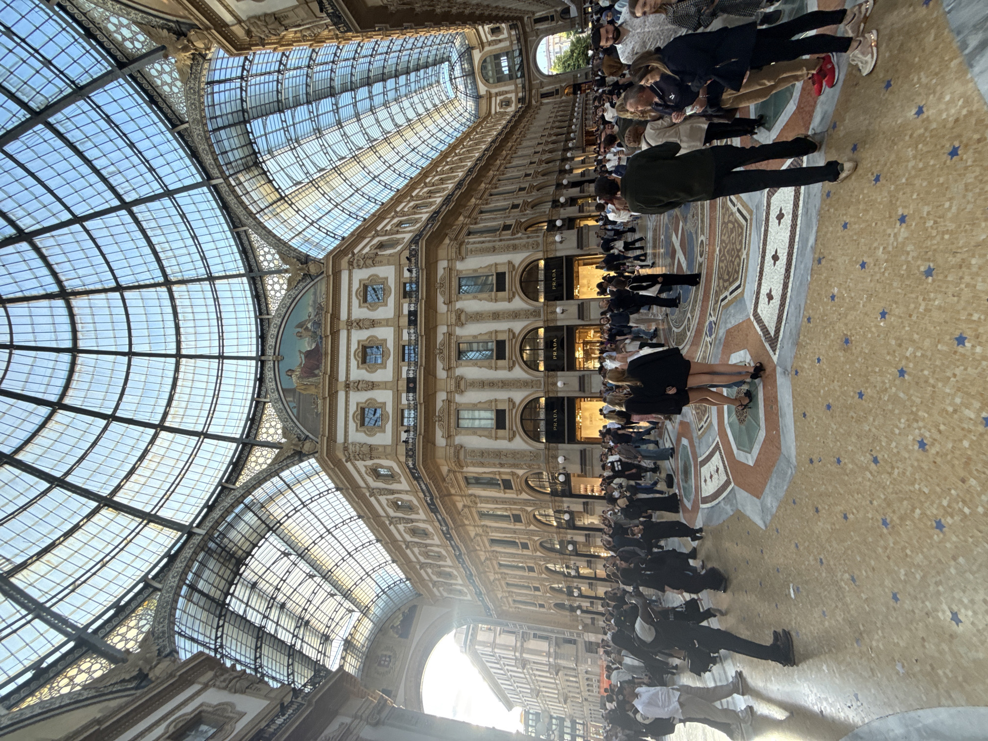 The image captures the Galleria Vittorio Emanuele II in Milan, Italy. The scene depicts a bustling interior with numerous people walking through the grand arcade. The architecture is characterized by its ornate detailing, including arched windows, intricate facade decorations, and a high glass dome. Luxury shops, including a Prada store, line the sides of the arcade. The floor is decorated with mosaics. The overall atmosphere is vibrant and touristy.