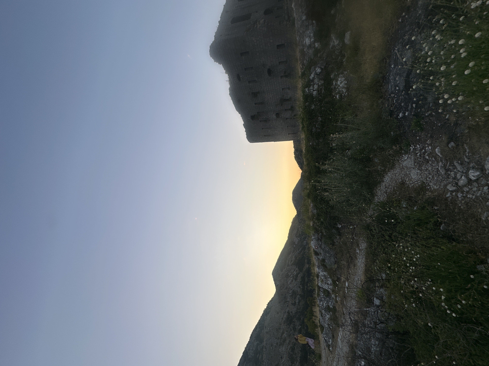The image depicts a remote, mountainous landscape at dusk, with a ruined stone fortress dominating the right side of the frame. A person wearing a yellow top and a pink skirt is visible in the distance on the left, standing on a rocky incline. The sky is a gradient of pale blue to soft orange from the setting sun. The terrain is rugged, with sparse vegetation and a dirt path winding through the foreground. The fortress appears to be an ancient structure, with weathered stone walls and numerous openings. The overall atmosphere is one of solitude and historical significance. The location is the San Giovanni Fortress in Brajici, Montenegro.