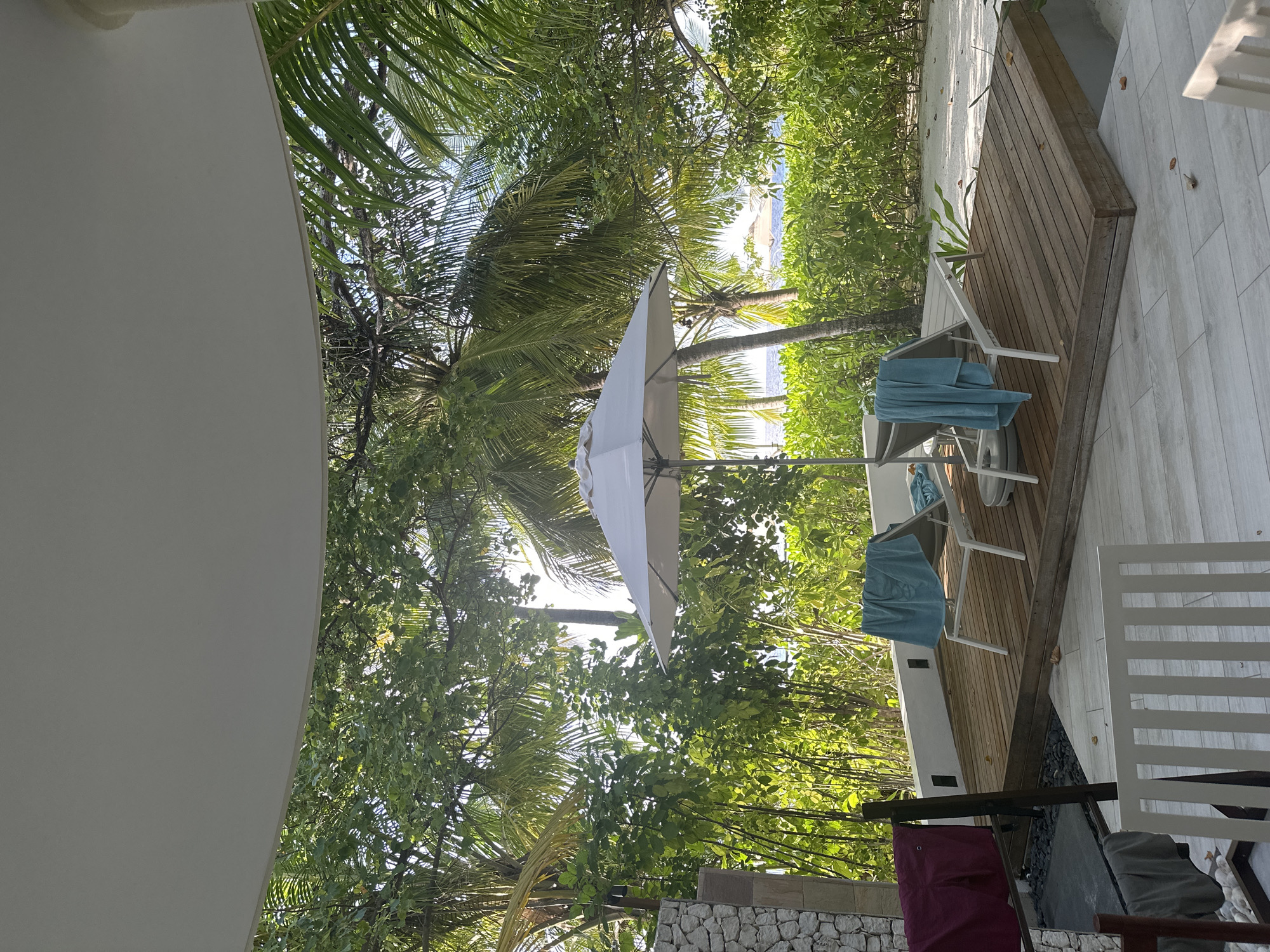 The image depicts a tropical resort scene in Halaveli, Maldives, featuring a wooden deck with two white lounge chairs. Each chair is draped with a blue towel. A white umbrella stands open between the chairs, providing shade. Lush green foliage, including palm trees, surrounds the deck. In the background, the ocean is visible with another umbrella on a distant beach. To the left of the deck, a stone structure stands next to a drying rack with several articles of clothing. The foreground shows a tiled patio area.