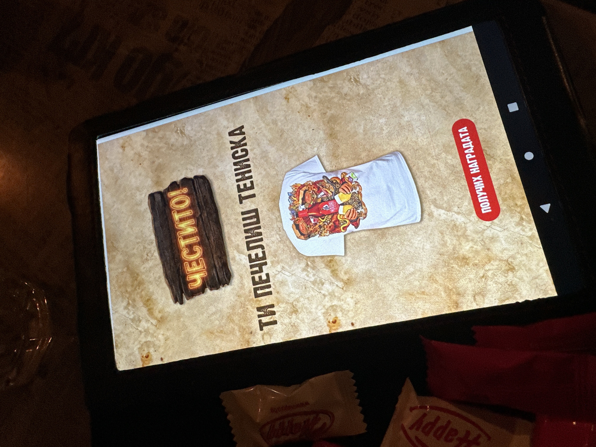 An overhead shot captures a black tablet displaying an interactive screen, resting on a dark, possibly wooden, surface. In the foreground, two wrapped candies are visible: one white wrapper with the word "Happy" (reversed) partially visible, and one red wrapper with "Addet" visible.
The tablet screen, illuminated against the darker surroundings, features a textured, light brown background resembling aged parchment. On the left side of the screen, a rustic, wooden sign-like graphic glows with the Cyrillic text "ЧЕСТИТО!" which translates to "Congratulations!". In the center of the screen, beneath a vertical line of Cyrillic text, lies a folded white t-shirt. The t-shirt is adorned with a vibrant, detailed graphic depicting various cartoonish food items like burgers, fries, and possibly drinks, along with other elements, suggesting a busy, celebratory design.
The vertical Cyrillic text next to the t-shirt reads "ТИ ПЕЧЕЛИШ ТЕНИСКА", meaning "YOU WIN A T-SHIRT". On the right side of the screen, a prominent red rectangular button with rounded corners displays the Cyrillic text "ПОЛУЧИХ НАГРАДАТА", which translates to "CLAIM PRIZE" or "I RECEIVED THE PRIZE". The overall scene suggests a digital promotion or game where the user has won a t-shirt, and the image captures the moment of revealing the prize and the prompt to claim it. The setting appears to be indoors, with the tablet screen being the primary light source in a dimly lit environment, possibly at night.