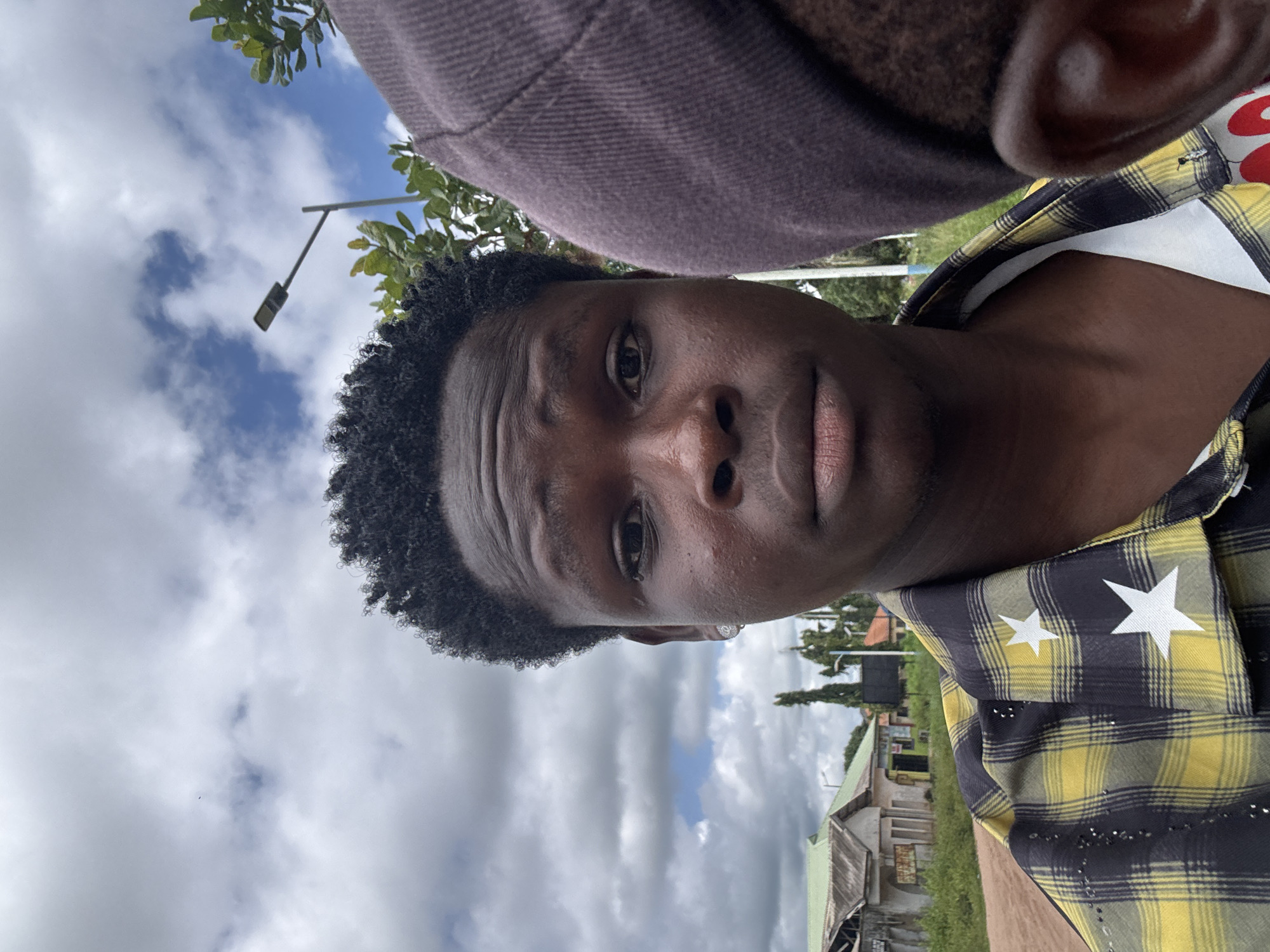 The image captures a medium shot of two individuals in what appears to be a semi-urban or rural setting in Ihiagwa, Nigeria. The primary subject, a young person with dark skin and tightly curled black hair, is positioned centrally in the frame, looking directly at the camera. They are wearing a yellow and black checkered shirt adorned with white stars. To the right of the primary subject, another person's head is visible, wearing a dark cap, their face obscured.
The background depicts a cloudy sky with patches of blue. A street lamp stands to the right of the primary subject, and behind them, a few buildings with green roofs and what seem to be shops or houses are visible, along with some vegetation and trees. The ground in front of the buildings is a dirt road. The overall scene suggests an outdoor environment under a somewhat overcast sky.
