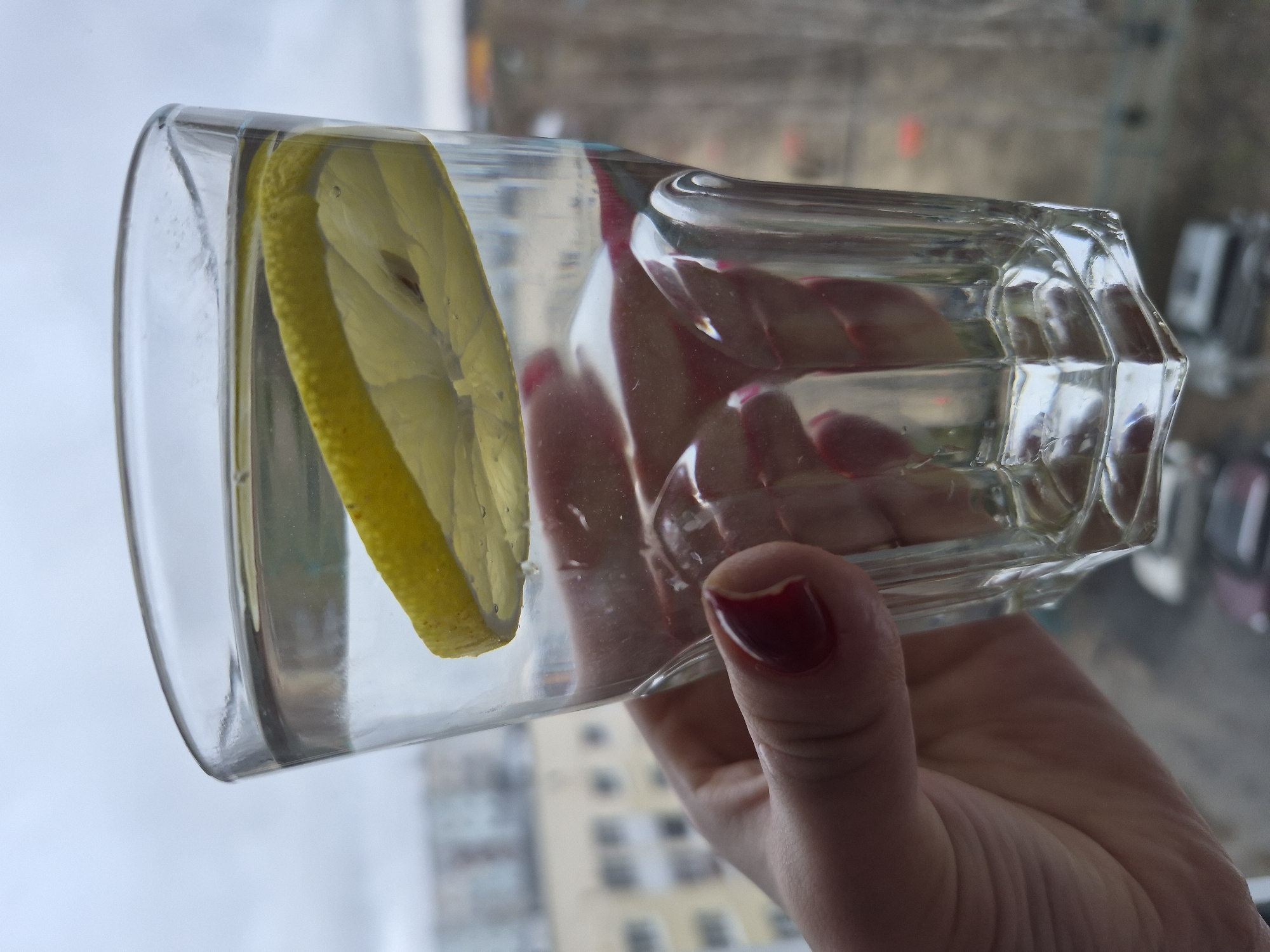 A hand with red nail polish holds a glass of clear liquid with a lemon slice. The glass is square with a round rim and faceted corners. The backdrop is an out-of-focus urban environment, with a residential building with windows visible. This scene is located in the city of Tobolsk, Russia.