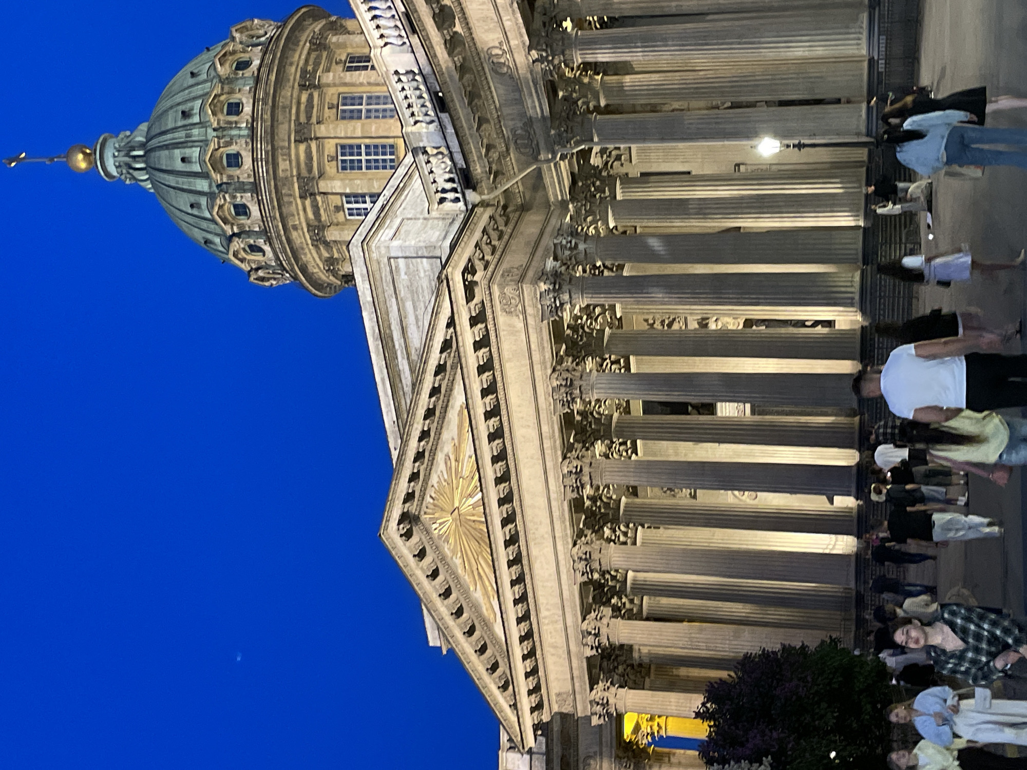 This image captures the Kazan Cathedral in Saint Petersburg, Russia, during the evening or twilight hours. The sky is a deep, uniform blue, indicating late day, while the cathedral's facade is illuminated by warm artificial lighting.
The central subject is the Kazan Cathedral, a large neoclassical structure featuring an extensive colonnade of numerous fluted Corinthian columns that support a prominent triangular pediment. The pediment's tympanum contains a golden, radiant symbol, often referred to as the Eye of Providence. Above the main structure, a large, patinated copper dome rises, topped with a golden cupola and a cross. The dome's drum features multiple arched windows.
In the foreground and mid-ground, a diverse group of casually dressed pedestrians occupies a wide, paved public square or walkway in front of the cathedral. People are seen walking, standing, and interacting in small groups. Notable individuals include a woman in a plaid shirt looking towards the camera in the left foreground, a woman in a light blue top and white skirt to her left, and a man in a white T-shirt with his back partially to the camera on the right.
To the left of the cathedral's columns, a dark green bush or small tree is visible, partially obscuring the lower part of the facade. Further left, partially visible behind the foliage and columns, is a vertical banner displaying distinct blue and yellow sections. On the right side of the paved area, a traditional-style streetlight stands illuminated, casting light on the surrounding ground and people. No specific dramatic actions or interactions are depicted; the scene portrays typical public activity around a prominent landmark.