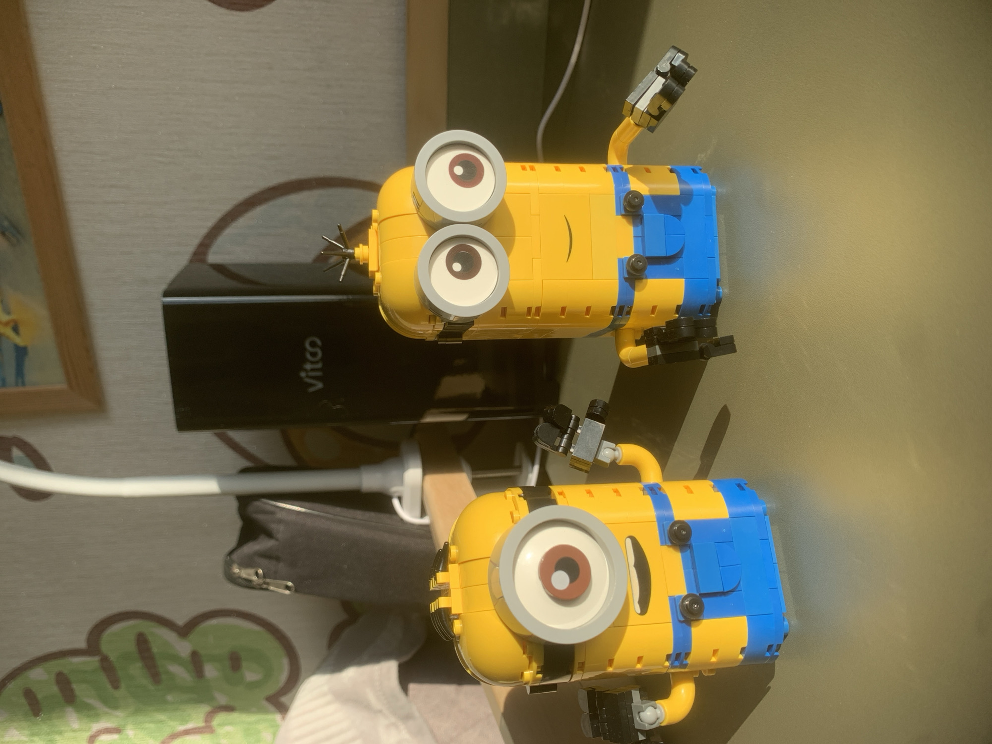 Two Lego minion figures are on a table. One has two eyes and the other one has one. A black power bank is between them. A white charging cable is connected to the power bank. A zipper bag is visible as well. A framed picture is on the side. The table has a patterned table cloth on one side and a light tan colored surface on the other. The location is not identifiable.
