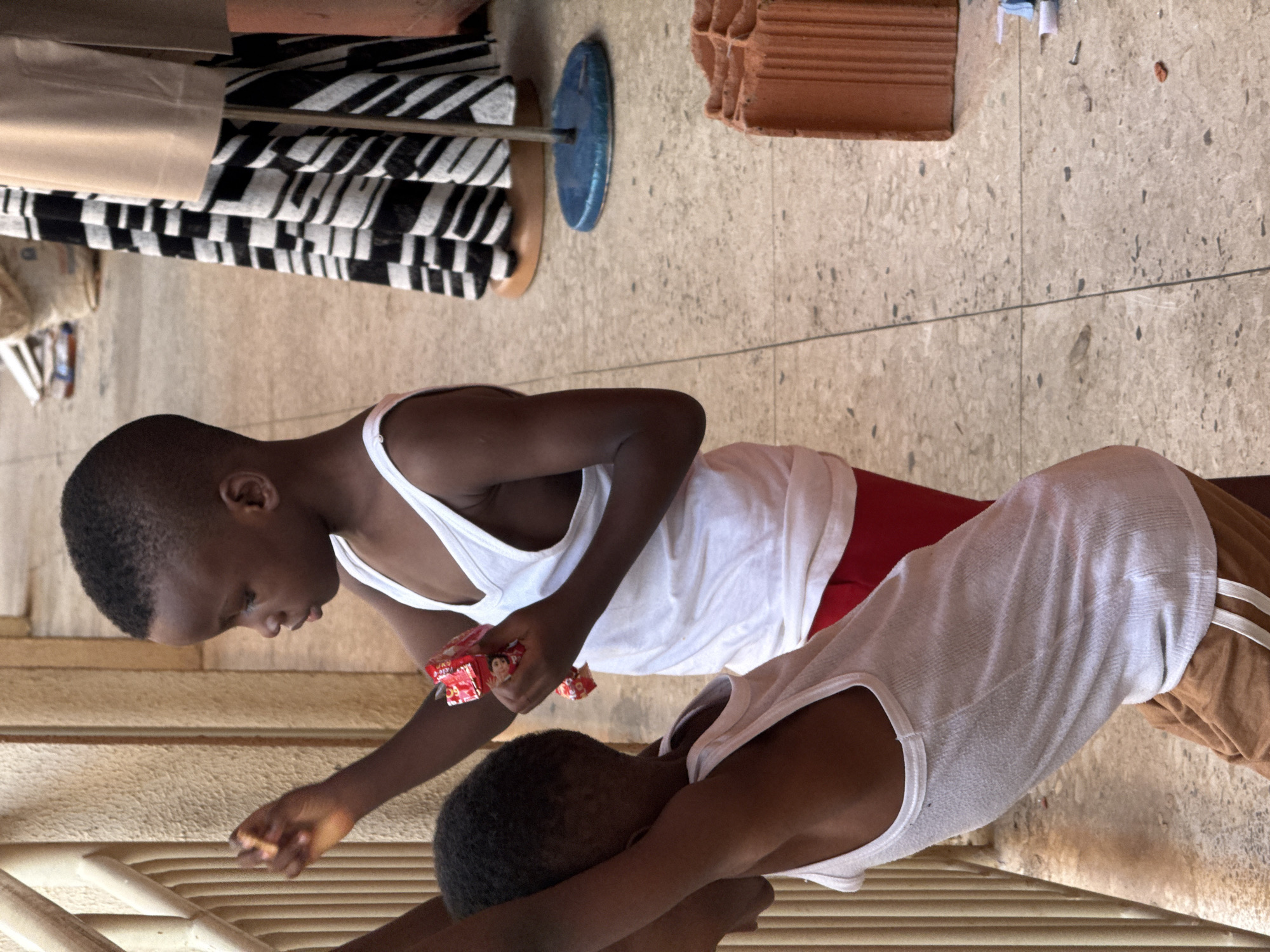 Two young dark-skinned children wearing white undershirts are interacting in an indoor or covered outdoor space. One child, closer to the camera, is kneeling, with arms crossed, the other is kneeling and holding a small red package with printed images. The floor is light-colored and textured. Behind the children are various objects including a stand with black and white striped fabric, a blue circle on a metal stand, and stacked red bricks. The location is identified as Abuja, Nigeria.