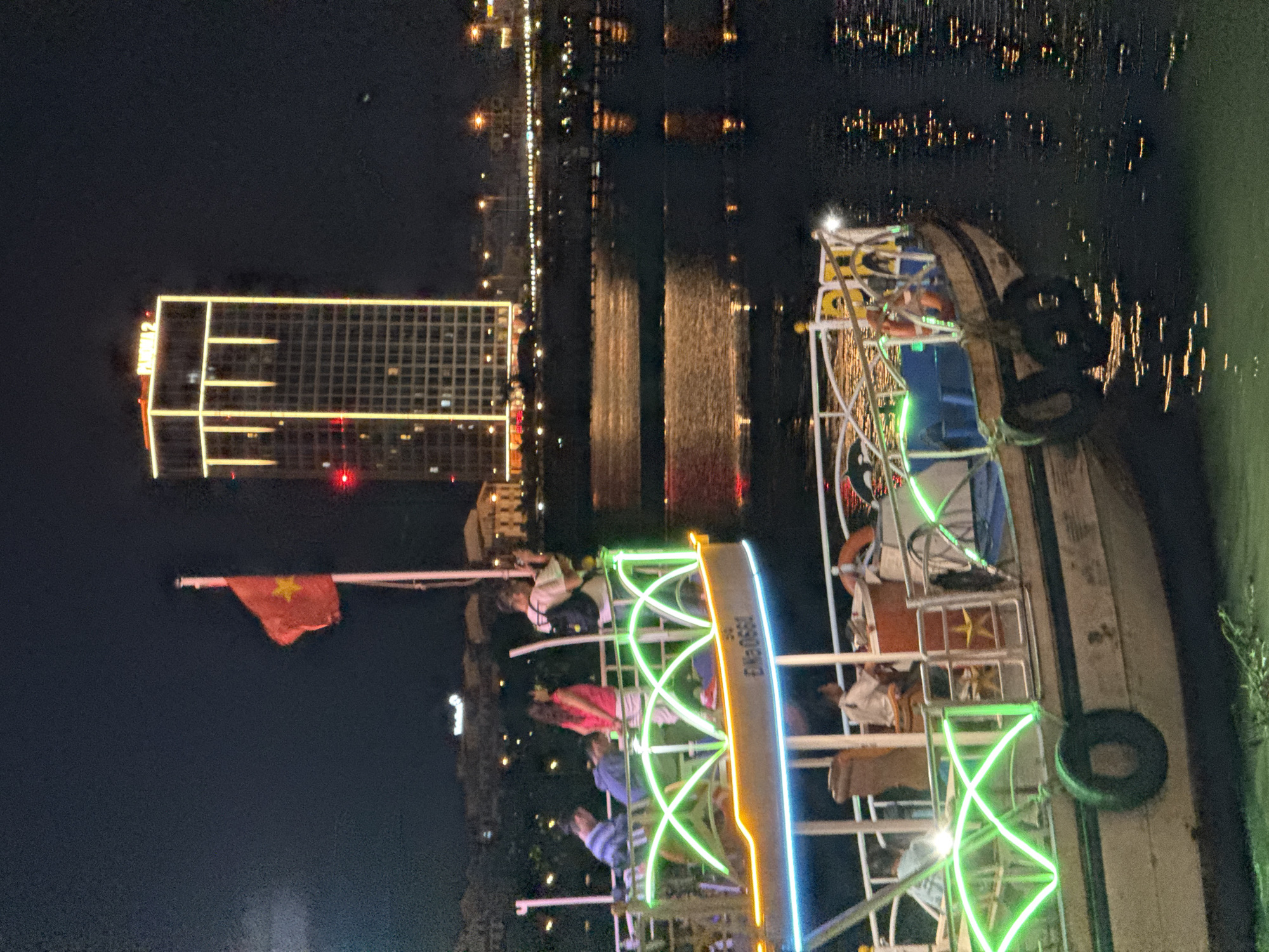 This media file presents a vibrant night scene from Da Nang, Vietnam, featuring a leisure boat navigating or docked on a river. The time of day is clearly night, evidenced by the dark sky and the extensive use of artificial lighting.
The central subject is a multi-deck boat, brightly illuminated with colorful neon lights in green, blue, and yellow, outlining its structure. Several individuals are visible on board, some standing and others seated, appearing to enjoy the night views. From a mast on the boat, a red flag with a yellow star, identifiable as the national flag of Vietnam, is flying. Visible text on the side of the boat reads "120800WA". An orange lifebuoy is also discernible on the boat's side.
In the background, a modern city skyline is illuminated. A prominent high-rise building on the left bank displays grid patterns of lighted windows and bright outlining, with the text "FLORA 2" visible on its upper section. Numerous other distant city lights twinkle, and their reflections shimmer across the dark river water, indicating a bustling urban environment. The activity taking place is likely a scenic night cruise or tour, offering passengers views of Da Nang's illuminated cityscape. The scene conveys a sense of peaceful enjoyment under the clear night sky.