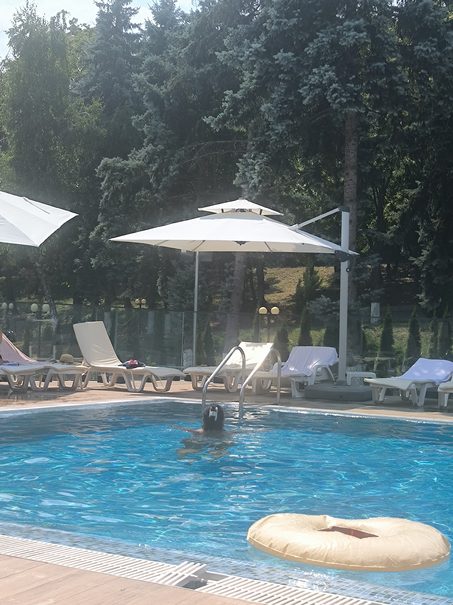 This is a medium shot taken outdoors of a swimming pool area, likely at a resort or hotel. In the foreground, a person is partially visible in the bright blue water, with their head and shoulders above the surface. A large, light-colored inflatable ring floats nearby in the pool. The poolside area features several white lounge chairs, some of which are covered with white towels, and large white umbrellas providing shade. Behind the pool, a glass fence separates the area from a lush green background of tall evergreen trees. The overall setting suggests a warm, sunny day in a tranquil outdoor environment. The location is specified as Ustia, Moldova.