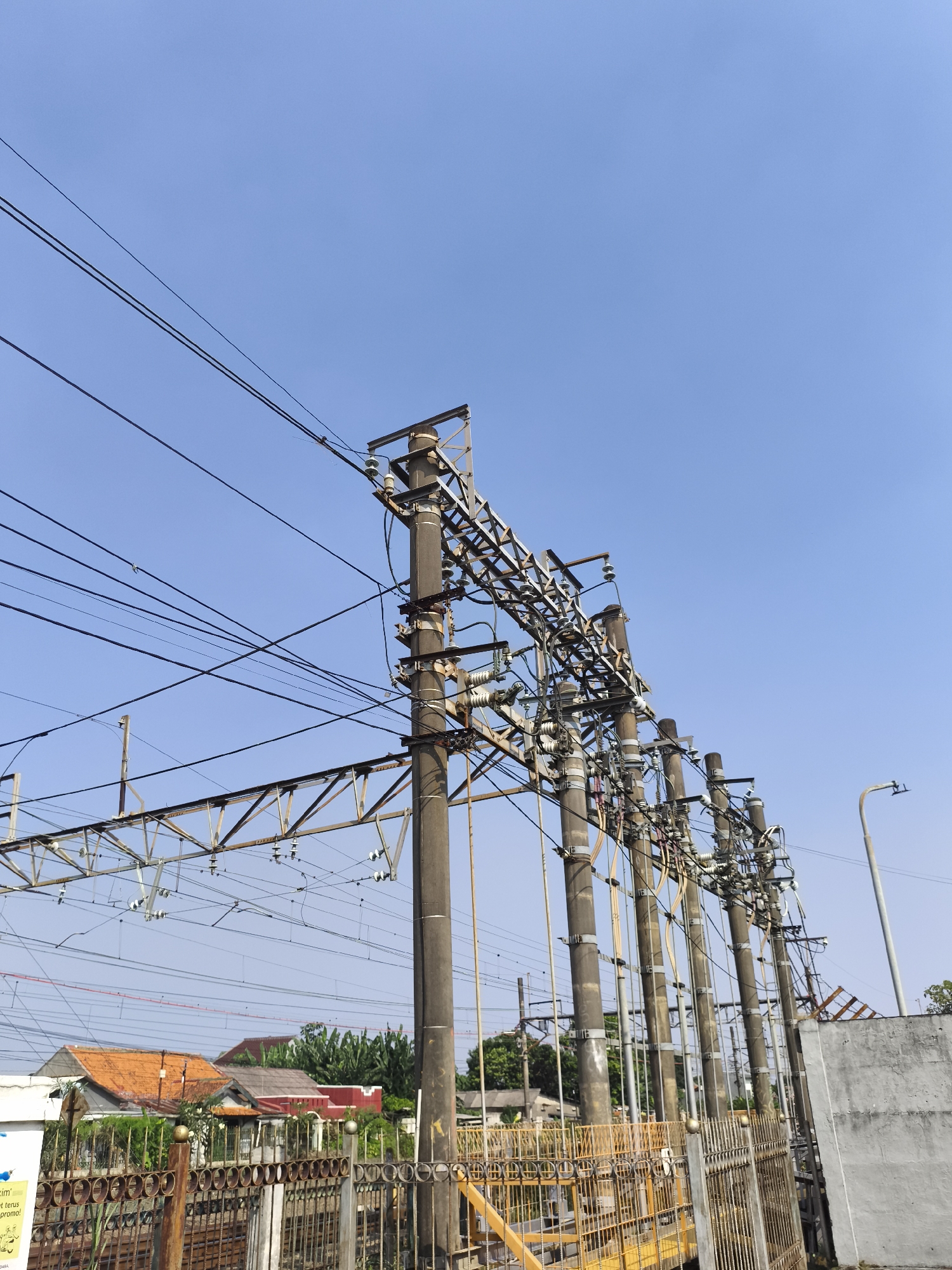 This detailed image captures an outdoor scene dominated by railway and electrical utility infrastructure, located within the city of Depok, Indonesia. The sky is a clear, bright blue, indicating fair weather and daytime conditions.
In the midground, a series of robust, grey concrete utility poles stand vertically, extending diagonally from the mid-left towards the right, receding into the distance. These poles are heavily laden with a dense network of electrical components, including numerous insulators, crossarms made of metal, and a multitude of thick black and lighter-colored electrical wires running horizontally, vertically, and diagonally between the poles and across the frame. These wires appear to be part of both a local power distribution system and the overhead catenary system for a railway line.
Intersecting the frame horizontally, a metallic lattice structure, characteristic of railway overhead line supports, is visible, extending across the upper-mid section. Below this, multiple parallel railway catenary wires are suspended.
In the lower foreground, a weathered metal fence with a decorative, somewhat rusty pattern is prominent, acting as a barrier. Parts of this fence, particularly towards the left, feature yellow accents. To the far right, a segment of a plain, light grey concrete wall is visible, along with a grey street lamp post extending upwards.
The background reveals a residential area with several houses. Their roofs display varying colors, including distinct orange/terracotta hues, grey, and red. Lush green trees and vegetation are interspersed among and behind these dwellings, suggesting a suburban or semi-urban environment typical of Depok. No individuals are visible in the scene.