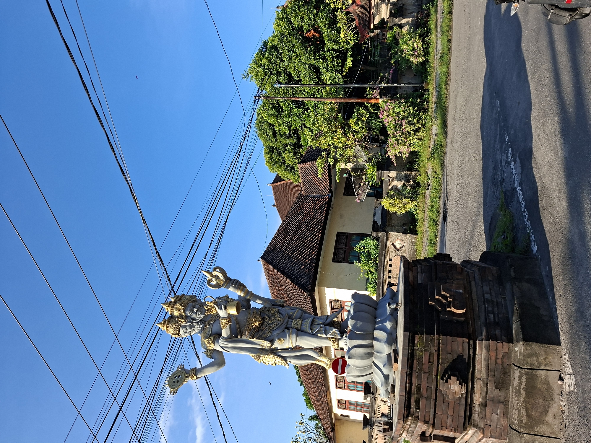 A statue of a multi-headed, four-armed deity stands on a lotus flower base, which is mounted on a brick pedestal. The statue is made of gray material with gold accents. It is positioned on the side of a road, with a house and lush green trees in the background. Overhead, a network of power lines crisscrosses the clear blue sky. The scene appears to be in Nusa Penida, Indonesia.