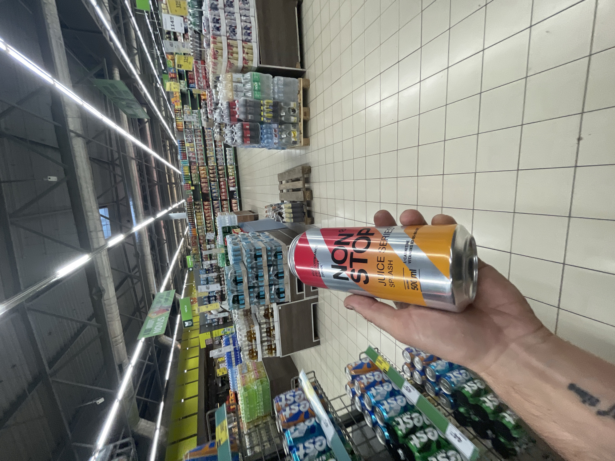 In the city of Vyshneve, Ukraine, a hand holds a 500ml can of "NON STOP" energy drink, with the label indicating "JUICE SERIES SPLASH". The can is predominantly silver with red and orange accents. The background reveals a brightly lit supermarket aisle, filled with various beverage products stacked on shelves and pallets. The flooring is a tiled pattern. In the distance, a green sign with the word "NOVUS" is visible, suggesting the supermarket chain. The perspective is from a low angle, looking forward and slightly upward.