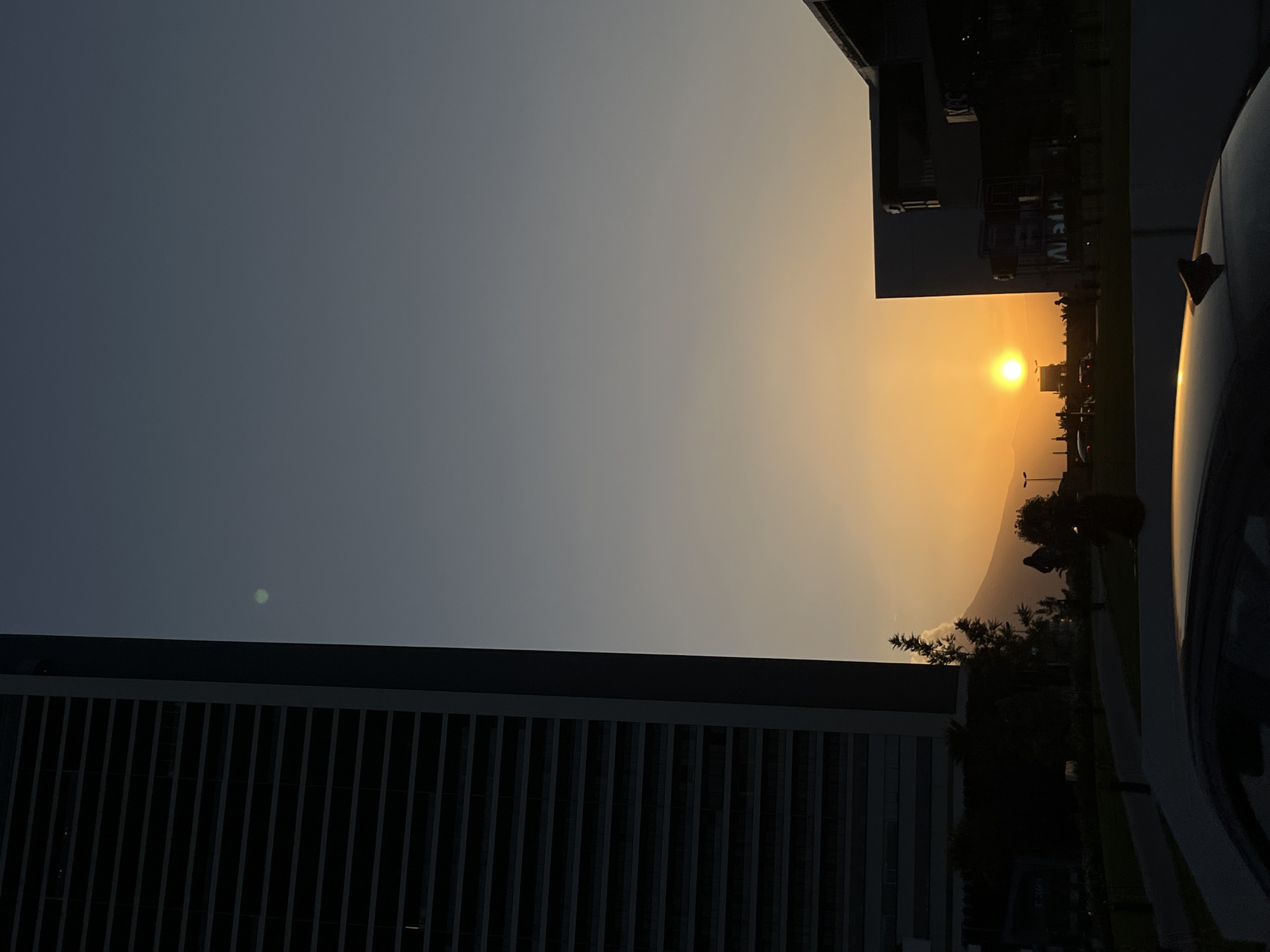 A striking sunset scene unfolds, viewed horizontally, with the sun positioned above a distant horizon. The sky exhibits a gradient from a deep, slate-gray in the upper left to a brilliant yellow-orange around the setting sun, which appears as a bright, circular orb.
In the foreground, a tall, modern building with distinct horizontal slats or louvers on its facade occupies the lower left portion of the image, appearing as a dark silhouette. On the right, the side and roof of a dark-colored car are visible, featuring a small "shark fin" antenna on its roof. Another large building, also silhouetted, is present in the upper right, behind the car.
The horizon line, visible beyond the buildings and car, is formed by silhouetted mountains or hills, further emphasizing the depth of the landscape. Dark trees and other vegetation are scattered between the foreground structures and the distant mountains. Faint, indistinct, and mirrored text, possibly beginning with "Wat" or similar, can be seen on a building in the mid-ground, likely a reflection. A small, circular, light green lens flare is also visible in the upper left sky.
The image captures the serene beauty of dusk, characterized by the warm glow of the sun against the darkening sky and the dramatic silhouettes it creates. No specific human activity is discernible, and the weather appears clear. The scene provides a glimpse into an urban or semi-urban environment, possibly in San Bernardino Tlaxcalancingo, Mexico, given the geographical context.