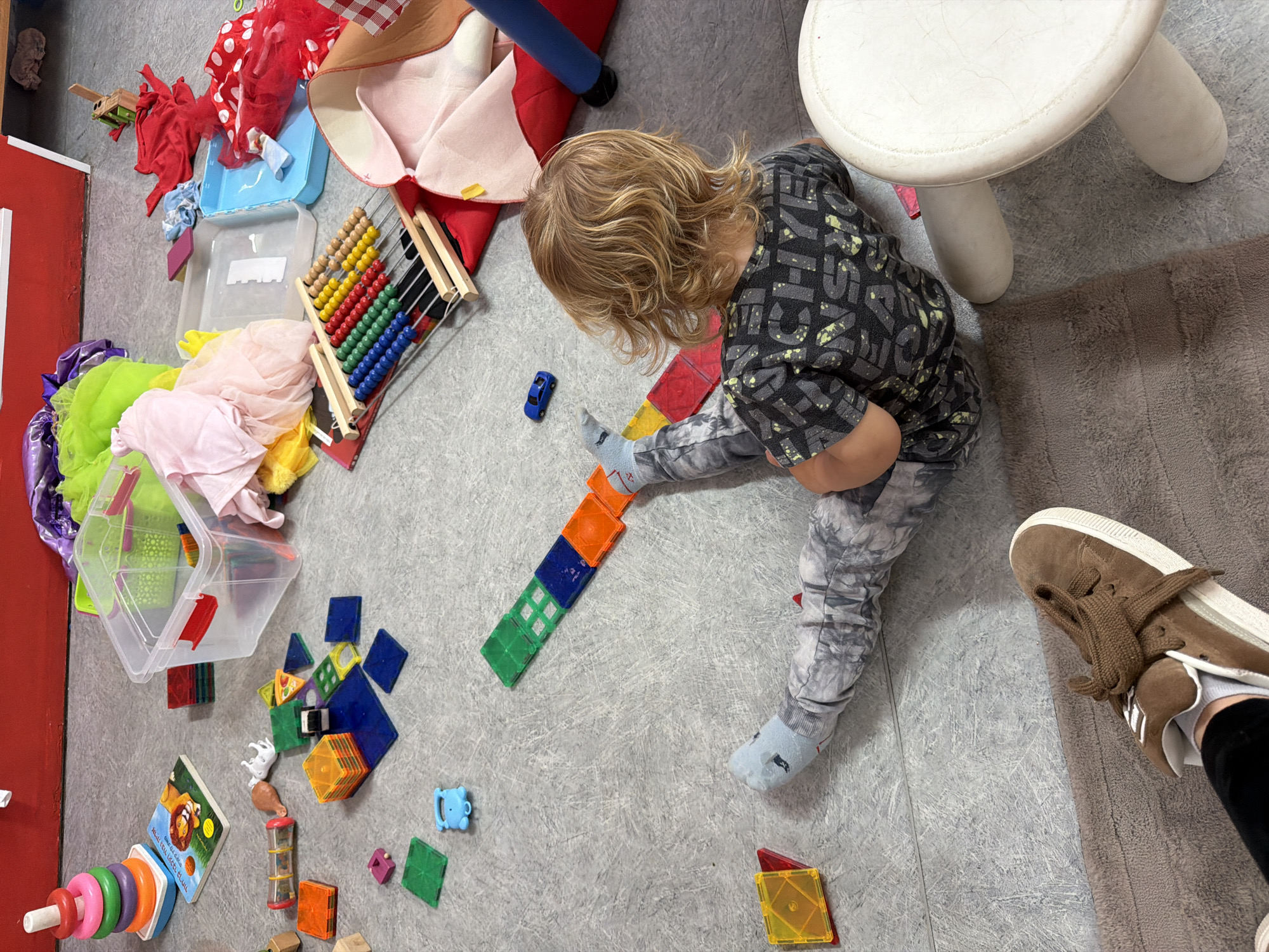 A child with blond, wavy hair is sitting on a gray, patterned floor, playing with colorful magnetic building blocks. The child is wearing a dark gray patterned shirt and gray tie-dye pants with light blue socks. The child is constructing a line of the blocks. An abacus, a book, stacking rings, and various other toys are scattered nearby. A small blue toy car is near the child. A small white stool is also present. In the background, there are fabric and storage containers. Part of a brown shoe is visible in the lower right corner. The location is Kiryat Bialik, Israel.