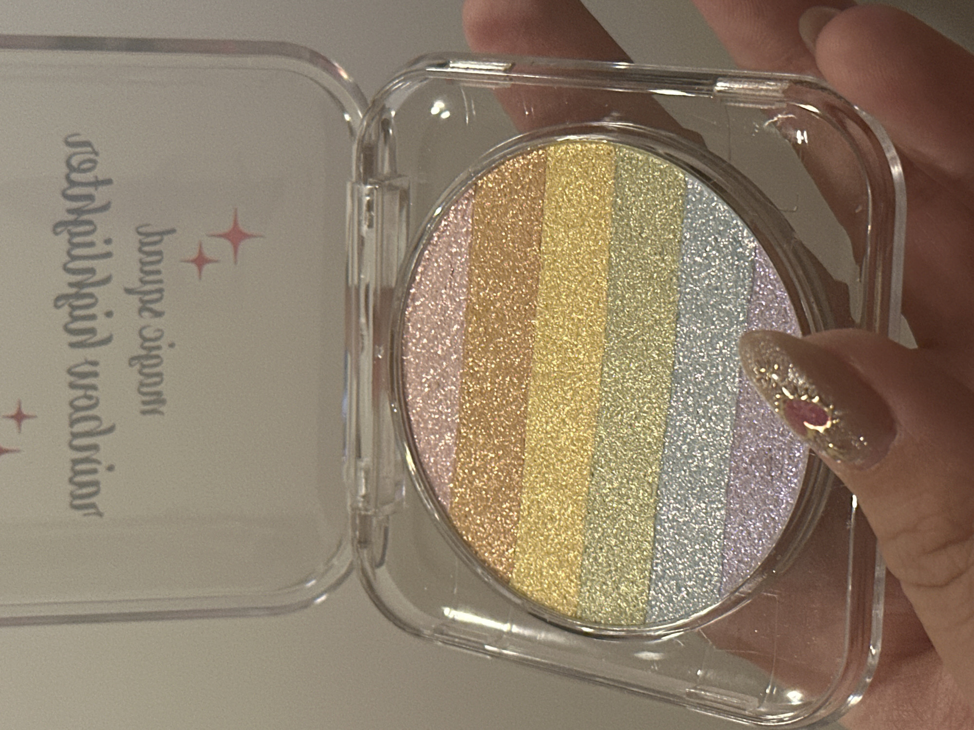 The image shows a person holding a square, transparent plastic compact containing a round, rainbow-striped powder makeup product. The compact is open. The colors in the powder are pink, gold, yellow, green, light blue and purple. A fingernail with glitter and a small heart embellishment is visible. The setting or location of the city of Zagreb in Croatia is not evident in the image.