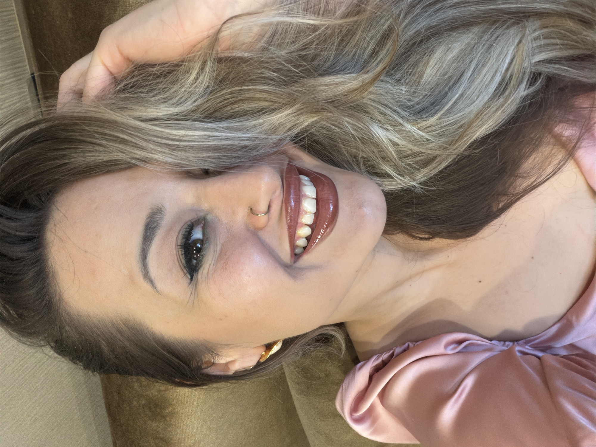 A woman lies on what appears to be a brown upholstered surface, possibly a couch or a bed. She is wearing a pink satin top. Her hair is light brown with blonde highlights, styled in waves, and her right hand is holding it. She is smiling, revealing her teeth, and has a nose ring. Her lips are glossy and a deep red-brown. The location and city of Krasnoyarsk, Russia are not determinable from the provided image.