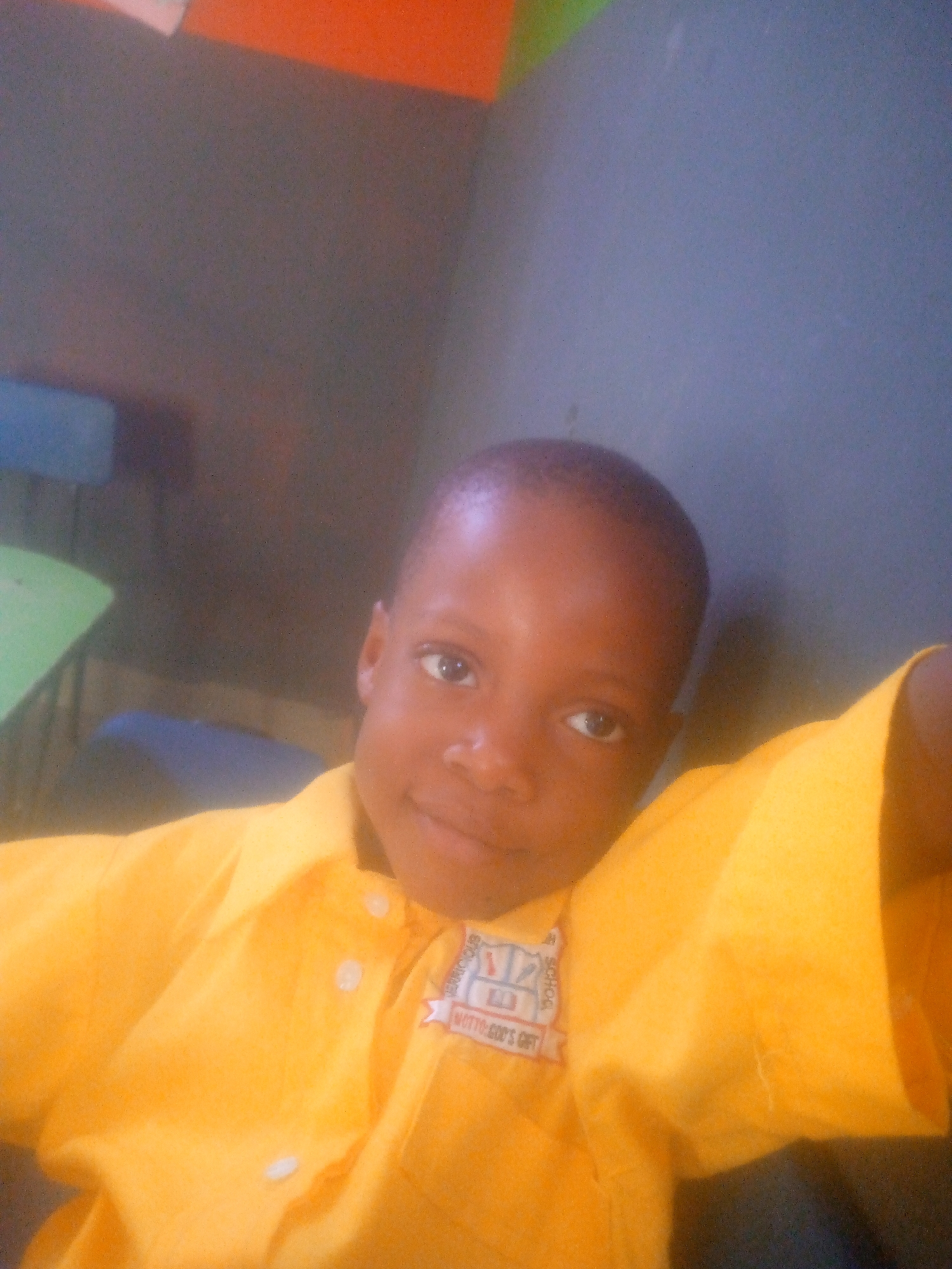 A young child wearing a yellow collared shirt with a school emblem is posing for a picture. The background appears to be an interior space, possibly a classroom, with a grey wall. A chair with a blue seat is slightly visible behind the child. Based on the text on the emblem, the child is associated with "Otto God's Gift" which is a school in Ikorodu, Nigeria.