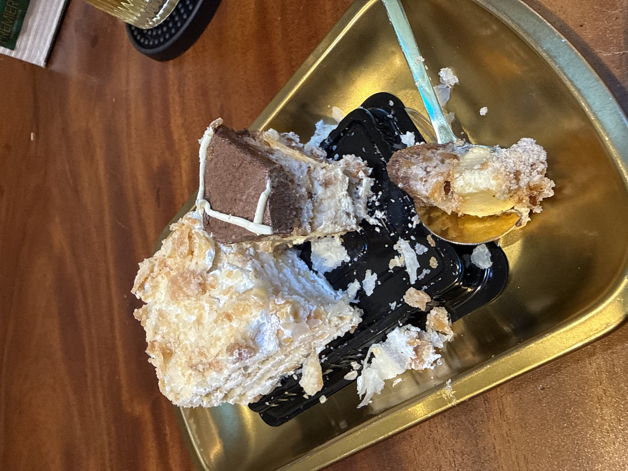 A close-up, eye-level shot shows a golden tray with two slices of Napoleon cake. The larger slice is on the left, and a smaller slice with chocolate ganache on top is to its right. Crumbles of cake and frosting are scattered across the tray. A spoon with a bite taken out of a piece of cake is in the lower right of the frame. The cake is on a black plastic container. The tray rests on a wooden table. In the background, a glass and a menu are visible. The setting appears to be a cafe or restaurant. The location of the cafe is specified as Bac Nha Trang Ward, Vietnam.