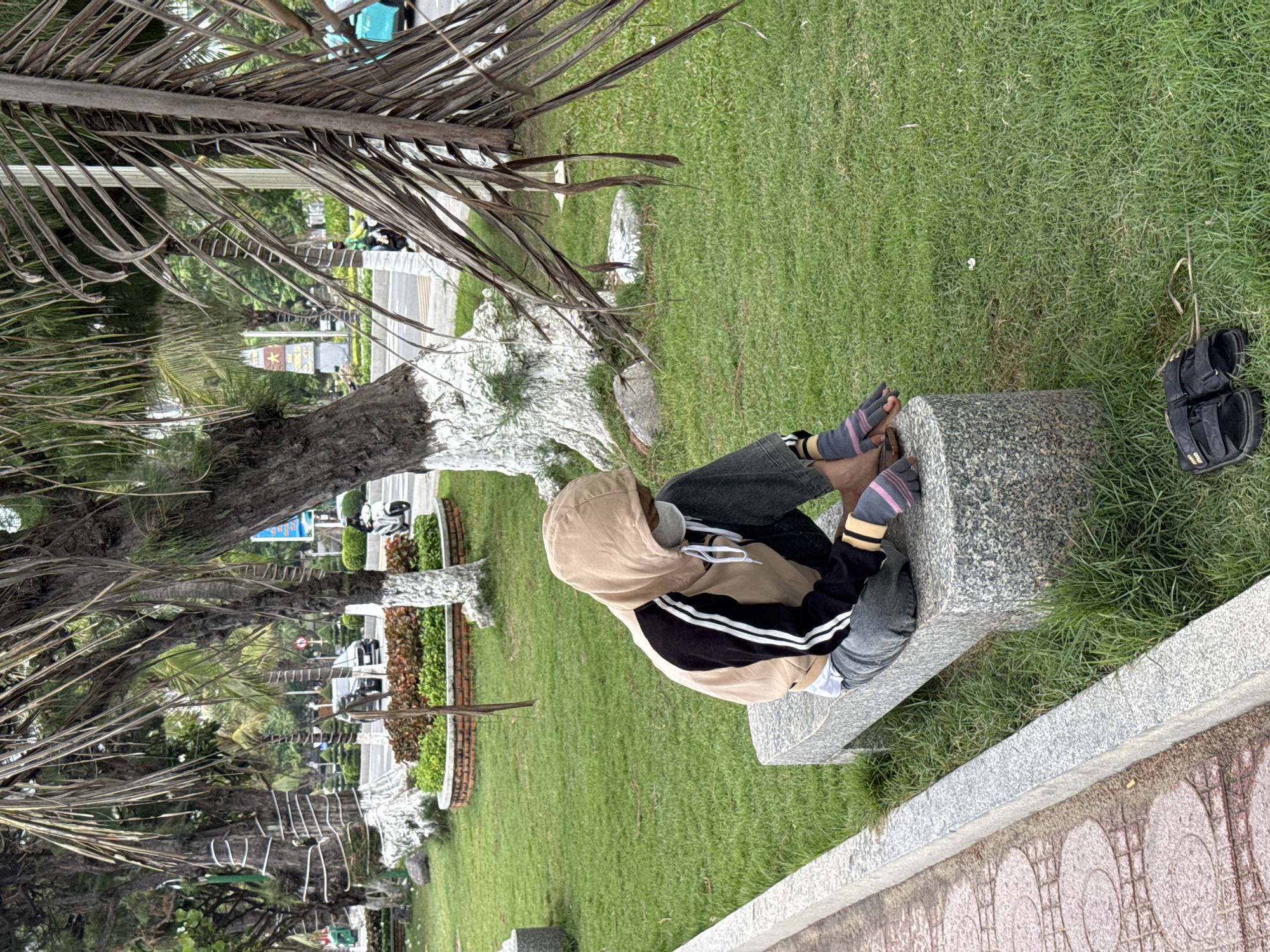 A lone individual wearing a face mask, a tan and black hooded jacket, and gray pants sits on a granite bench in a park in Nha Trang, Vietnam. The individual is wearing fingerless gloves with pink stripes. To the right of the bench, a pair of black sandals rests on the grass. The park features a sidewalk with a decorative pattern, lush green grass, and several trees, including palm trees with dry fronds. In the background, traffic can be seen on a road, including cars, scooters, and a turquoise vehicle. A monument with a red star is visible further in the background.