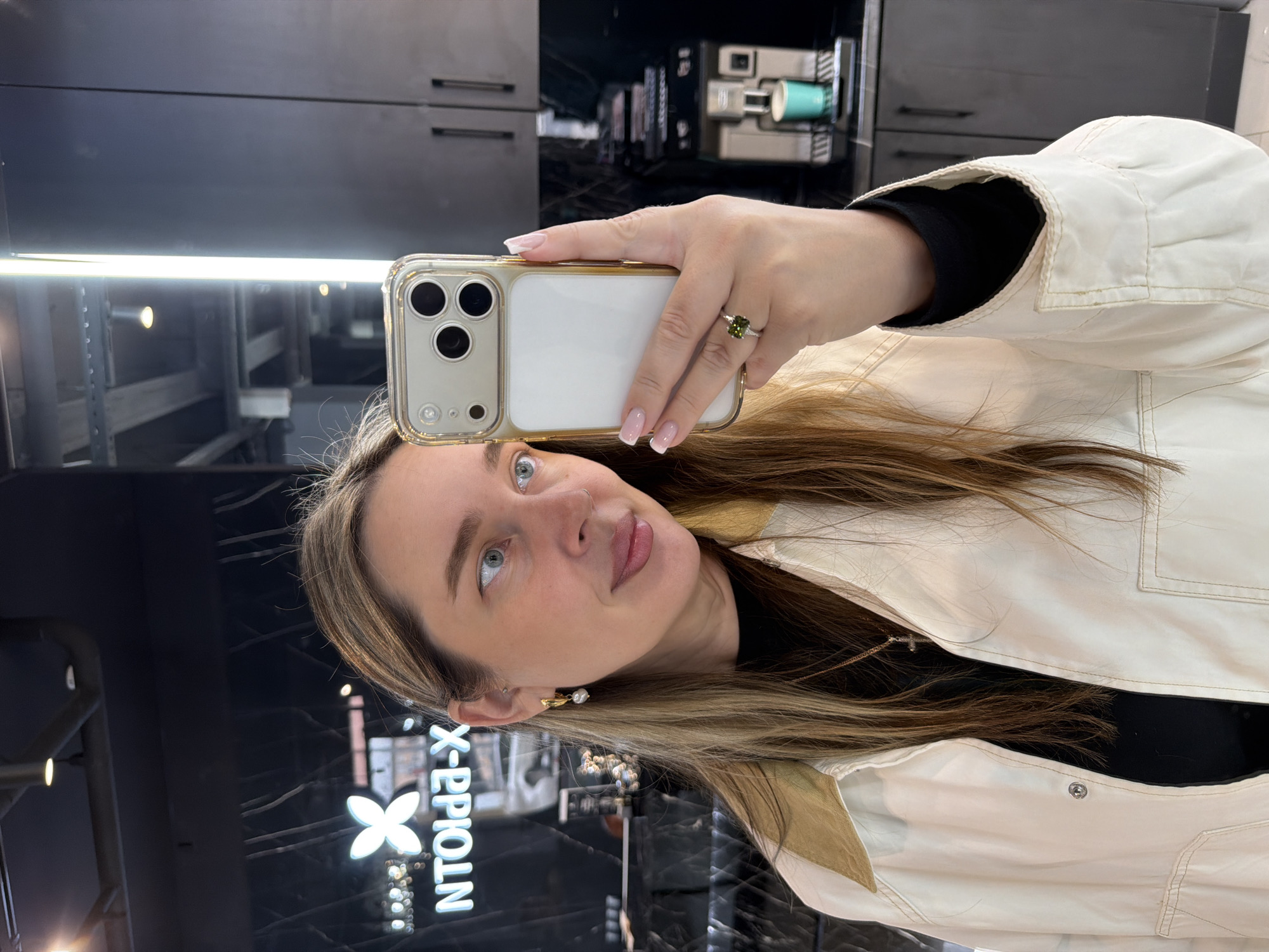 A young woman is depicted taking a selfie in what appears to be a mirror or reflective surface in an indoor setting. She is oriented with her head slightly tilted and her gaze directed upwards and to the left, away from the phone camera. She has long, light brown hair with blonde highlights and a gentle smile. Her eyes are blue or grey.
She is wearing a cream or off-white collared jacket over a dark top, with a thin necklace featuring a small cross pendant visible. On her left ear, she has a gold-toned earring with a white pearl. Her right hand, holding a smartphone in a clear case, is prominent in the foreground, showcasing her neatly manicured nails with French tips and a ring with a large green gemstone on her ring finger. The smartphone itself has a multi-lens camera system, indicative of a modern model.
The background, seen in the reflection, is dark and modern. It includes black cabinets or shelving units, and bright linear lighting fixtures are visible. In the upper right corner of the reflection, a coffee machine with a teal cup is discernible. To the left of the woman's head in the reflection, a lighted sign with a stylized floral or star emblem is visible, displaying reversed text that, when read correctly, spells "NTOIPP-X". The overall scene suggests a contemporary commercial or lounge environment.