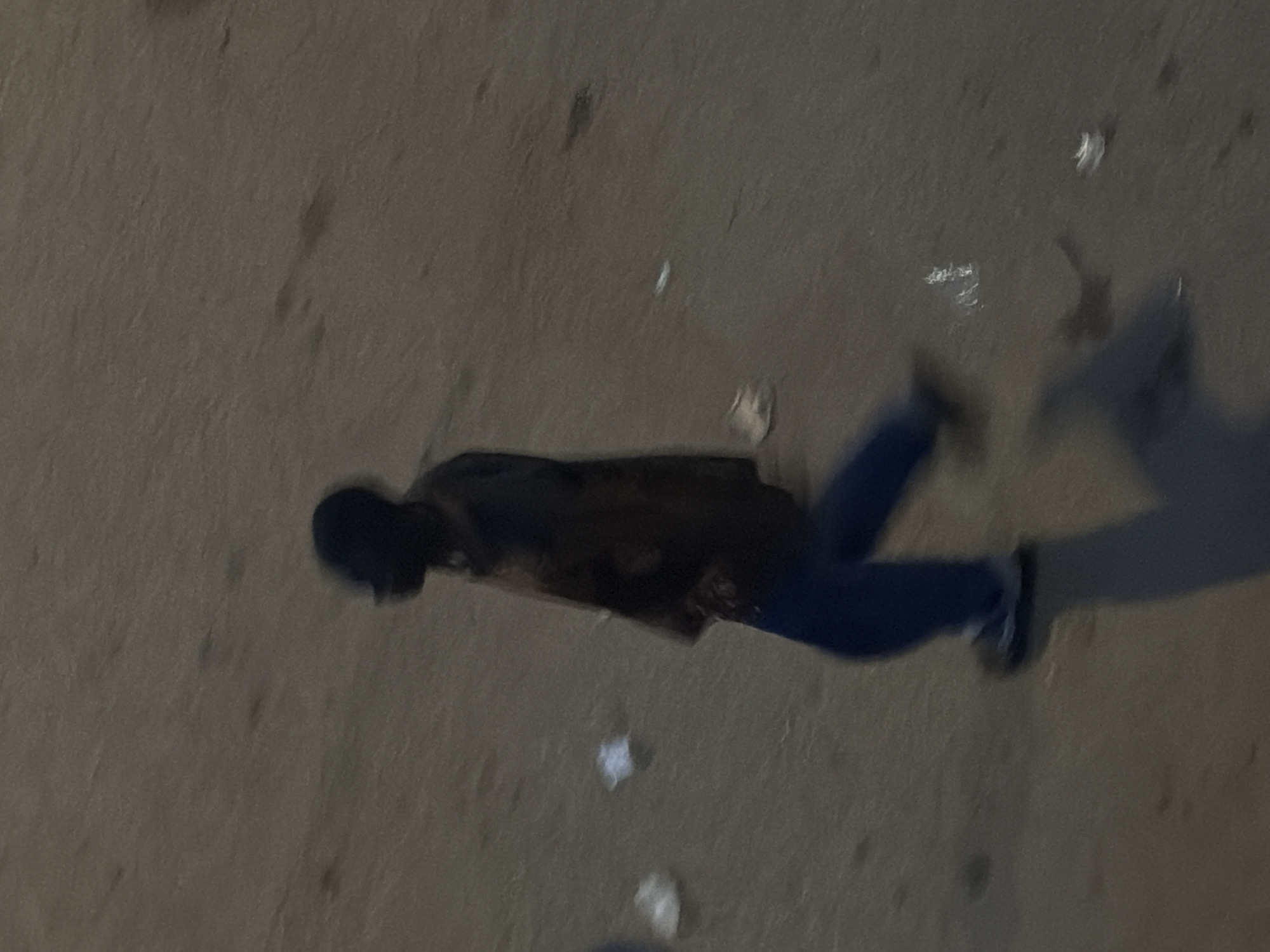 An out-of-focus, medium-angle shot captures a blurred silhouette of a person walking on a sandy or dusty surface. The person, viewed from the side, appears to be a male wearing a dark top and blue trousers. Their feet are clad in dark shoes with white accents. The ground is a uniform brown, with several small, white pieces of debris scattered across it, potentially litter or natural detritus. The lighting is dim, creating a shadowed environment and contributing to the overall blurriness of the image. The scene is likely outdoors, on a street or pathway in Bukuru, Nigeria, given the context.