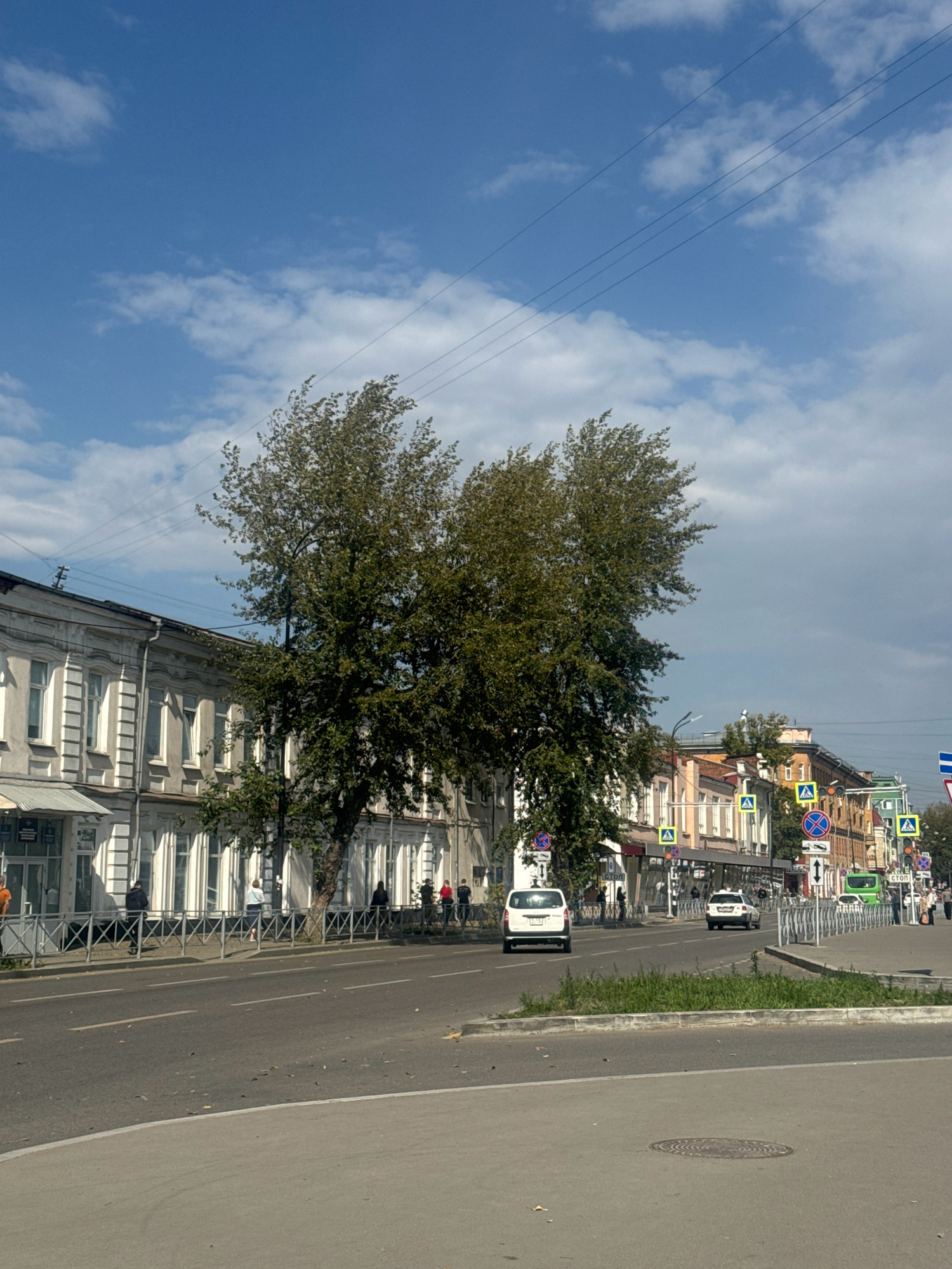 The image captures a street scene in Irkutsk, Russia, on a bright, cloudy day. In the foreground, a paved road curves to the left, with a grey concrete barrier and a patch of green grass separating it from the sidewalk. The sidewalk is lined with a metal fence, behind which are pedestrians walking along a row of buildings.
The buildings, constructed in a classical style with light-colored facades and numerous windows, appear to be old and historic. Several trees with dense green foliage are situated along the sidewalk, partially obscuring the buildings.
Further down the street, more buildings of varying architectural styles and colors are visible, interspersed with traffic signs and streetlights. Two white cars are driving on the road, one closer to the viewer and another further away. A green bus is also seen in the distance. The sky is a vibrant blue with scattered white clouds, and power lines stretch across the upper portion of the frame. The overall impression is that of a typical urban street in a historic Russian city.