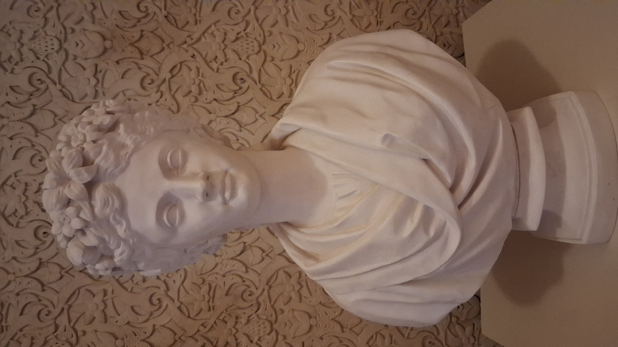 A white marble bust of a young woman is positioned centrally in the frame, facing forward. She has an elaborate floral headdress, curly hair, and is draped in flowing garments. The bust rests on a tiered white pedestal. The background is a beige, textured wall with an ornate, repeating pattern. The image appears to be taken indoors, possibly in a museum or historical site, with soft lighting. The location is the National Palace of Sintra, in Sintra (Santa Maria e São Miguel, São Martinho e São Pedro de Penaferrim), Portugal.