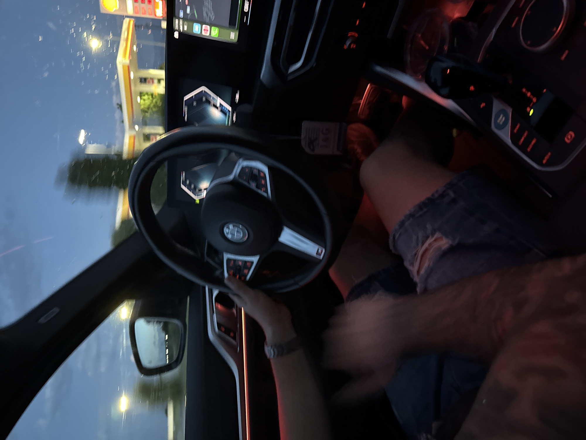 The photo is taken from inside a car, presumably stopped at a Shell gas station. The steering wheel is visible, with a hand interacting with it. The driver is wearing distressed denim shorts. The background shows a Shell gas station visible through the windshield. The image suggests an interior view of a car parked at a gas station at dusk or dawn, with the driver present. The location is not readily discernible as Dragor, Bulgaria.
