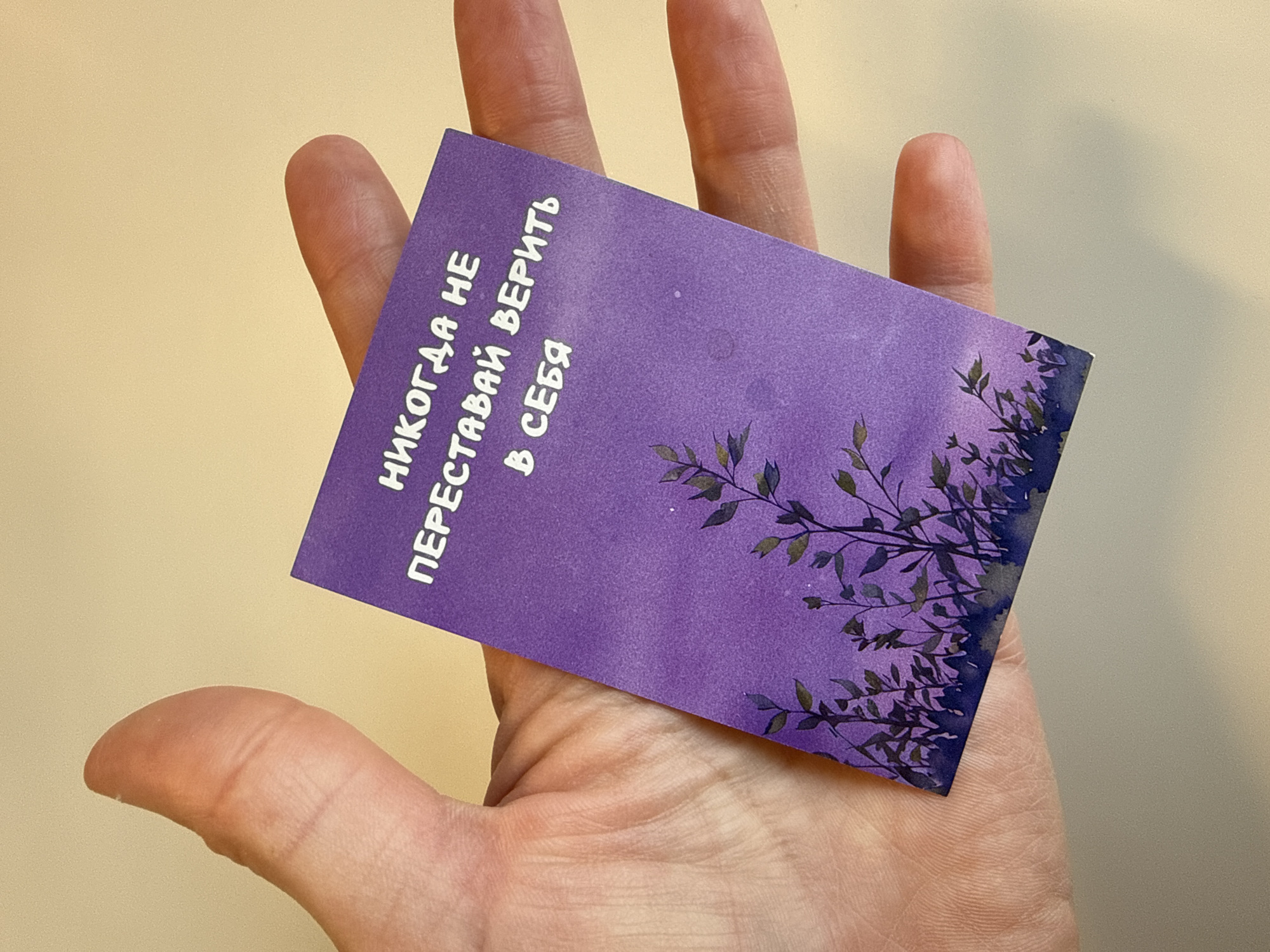 A hand is holding a small, purple card with white lettering in Russian. The text says "Never stop believing in yourself". There is a design of dark, leafy branches along the right edge of the card. The background is a plain, light yellow wall. The image was taken indoors.
