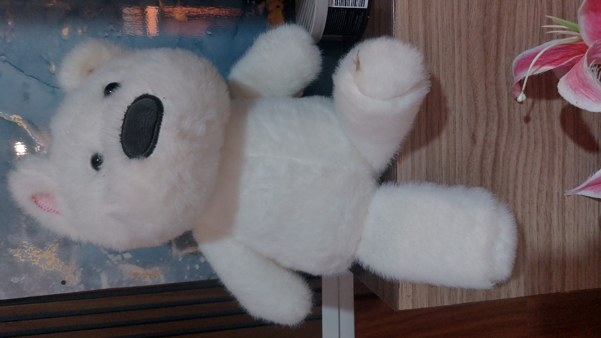 A white, plush teddy bear is sitting on a wooden surface. The teddy bear has black eyes and a black nose. Its ears are visible and appear to be slightly pink on the inside. The background behind the teddy bear is a wall with a blue and gold abstract pattern and some vertical dark lines on the left side. In the foreground, part of a pink and white lily is visible. The scene appears to be indoors, possibly in a residential setting. There is no indication of the city of Medan, Indonesia in the image itself.