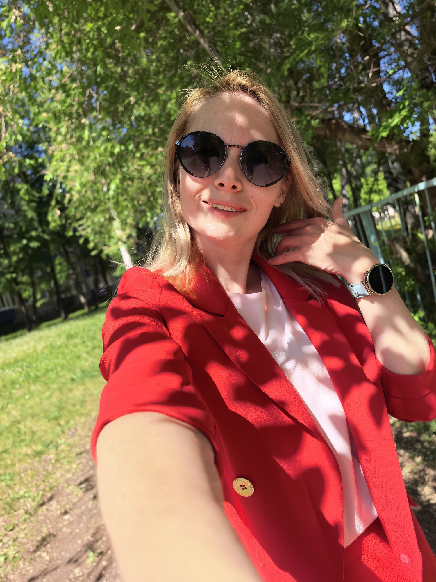 A light-skinned woman with blonde hair and dark sunglasses is taking a selfie outdoors on a sunny day. She is wearing a bright red blazer over a light pink top. The woman's left hand is raised to her hair, and a smartwatch is visible on her left wrist.
In the background, there are trees, grass, and a fence, suggesting a park or green space. The woman is standing on a path or walkway. According to EXIF data, the location is Sterlitamak, Russia, suggesting this photo was taken there.