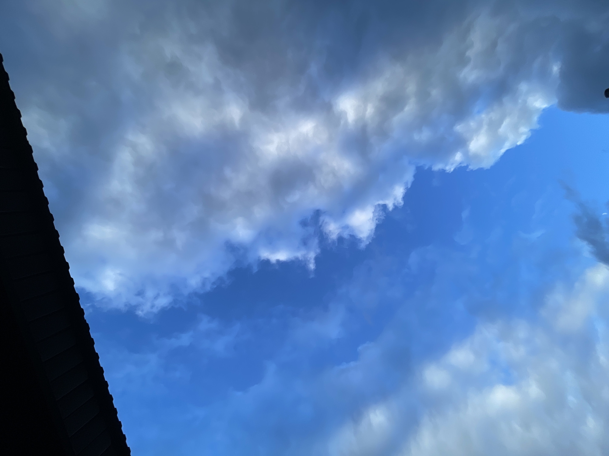 The image shows a dramatic sky filled with dark, brooding clouds and patches of bright blue sky. The clouds appear heavy and textured, suggesting the possibility of rain or a storm. A dark, angular object, likely a roofline of a building, occupies the lower left portion of the frame, contrasting sharply with the expansive sky. The lighting suggests it is daytime, possibly late afternoon or early evening, given the intensity of the cloud formations and the deep blue of the clear sky. The scene evokes a sense of anticipation, perhaps even a touch of foreboding, due to the ominous appearance of the clouds. No people or text are visible in the image. The setting appears to be outdoors, with the perspective looking upwards towards the sky.