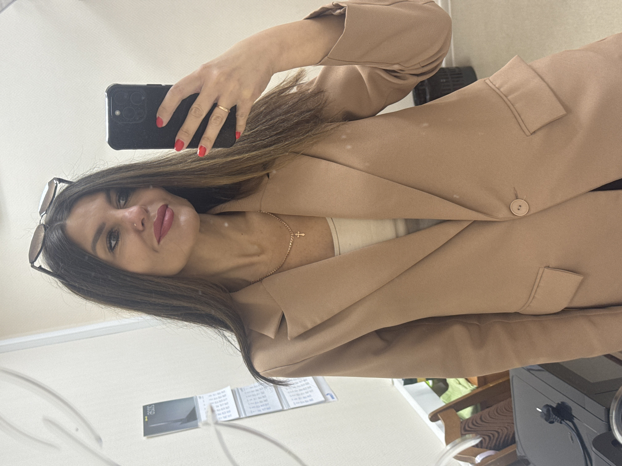 A woman in a beige blazer and a light-colored top is taking a mirror selfie. She has long, dark hair and is wearing red lipstick and a gold cross necklace. A pair of sunglasses is perched on her head. Her right hand, with red-painted nails, holds a black smartphone, and she wears a thin gold ring on her left ring finger. In the background, a wall with a textured pattern is visible, along with a calendar hanging on the left. A glimpse of office furniture, including chairs, a desk, and a trash can, can be seen. The overall setting appears to be an office or a similar indoor workspace, potentially located in the city of Chaikovsky, Russia.
