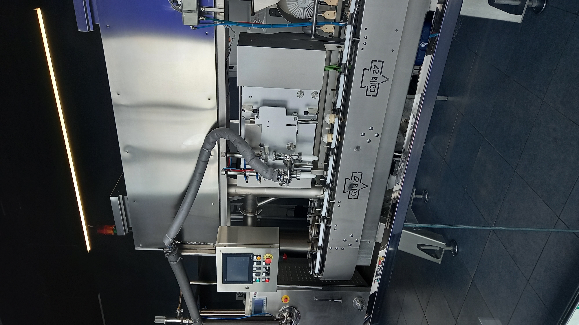 An industrial ice cream filling machine is shown in operation, likely in a food production facility. The machine is predominantly made of stainless steel and features a conveyor belt system that moves small ice cream cones. Above the conveyor, a filling mechanism with multiple nozzles is dispensing what appears to be ice cream into the cones. A control panel with a touchscreen interface and various buttons is visible on the left side of the machine. The environment appears to be clean and sterile, with tiled flooring and industrial lighting overhead. The overall scene depicts a high-tech manufacturing process for frozen desserts.