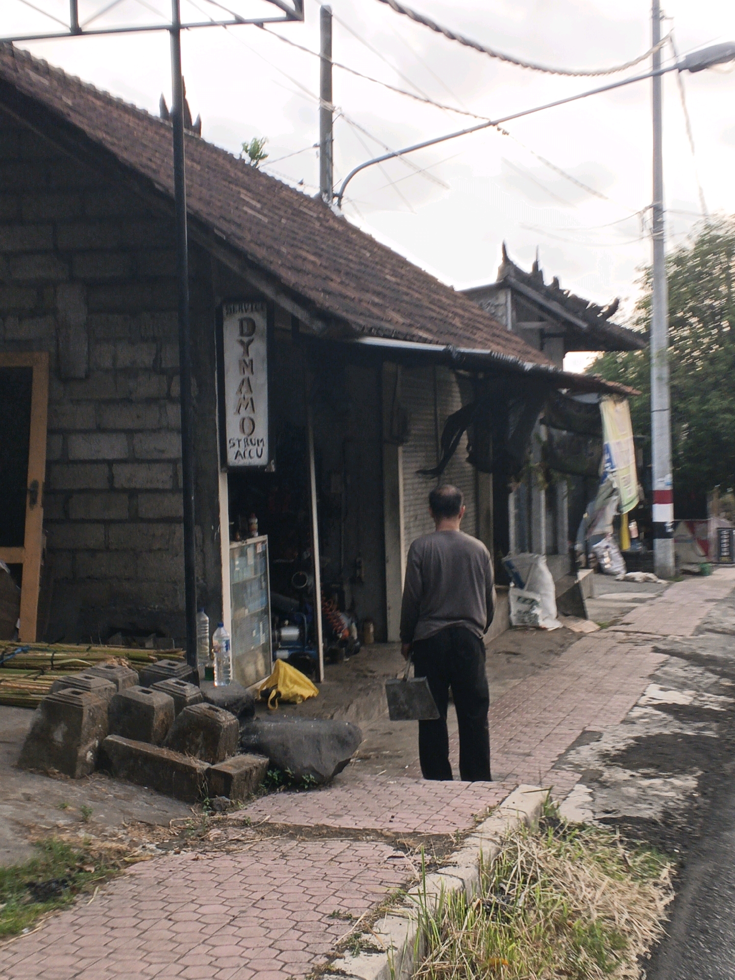 A medium shot captures a man walking away from the camera down a paved walkway in Sukawati, Indonesia. The man, wearing a grey long-sleeved shirt and black pants, carries a dark square bag in his right hand. The walkway is bordered by a grassy area and a dirt path.
To the left of the walkway is a single-story building constructed of grey cinder blocks with a red tile roof. A sign hanging from the building reads "SERVICE DYNAMO STRUM ACCU." In front of the building, there is a pile of square stone blocks and some bamboo poles. A yellow bag lies on the ground near the building's entrance.
The street itself is paved and appears wet in places. To the right of the walkway, a utility pole with wires runs vertically. The sky is overcast. The scene depicts a street in a commercial area, likely a workshop or service center.