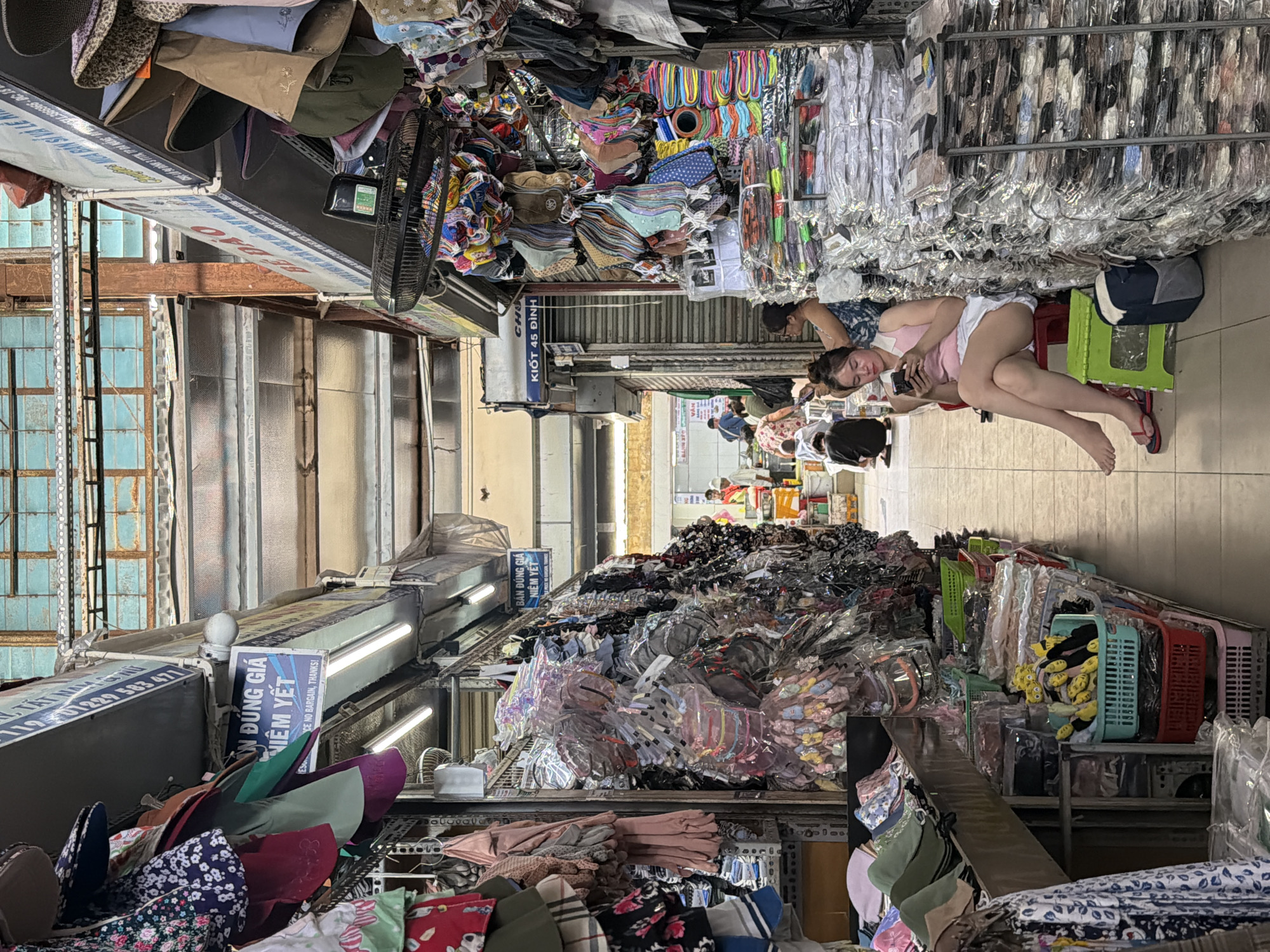 This image depicts a bustling indoor market scene, likely in Đà Nẵng, Vietnam. The aisle is densely packed with a variety of goods, including clothing, accessories, and other merchandise. The items are piled high on shelves and racks on both sides of the corridor, creating a cluttered yet vibrant atmosphere. A woman in a pink tank top and white shorts sits on a plastic stool in the aisle, looking at her phone. Further down the aisle, other people are visible, moving through the market. Signs in Vietnamese indicate fixed prices and discourage bargaining. The scene conveys a sense of activity and commerce within a traditional market setting.