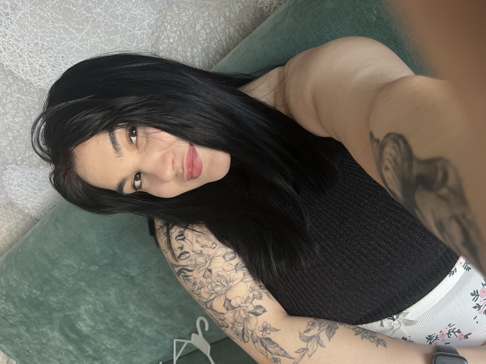 A woman with long, black hair is lying on a green cushion or couch. She is wearing a black, sleeveless top and white bottoms with a floral print. Tattoos are visible on both arms, including floral designs and a face on her right arm. The background includes a textured white wall. The location is Chaikovsky, Russia.