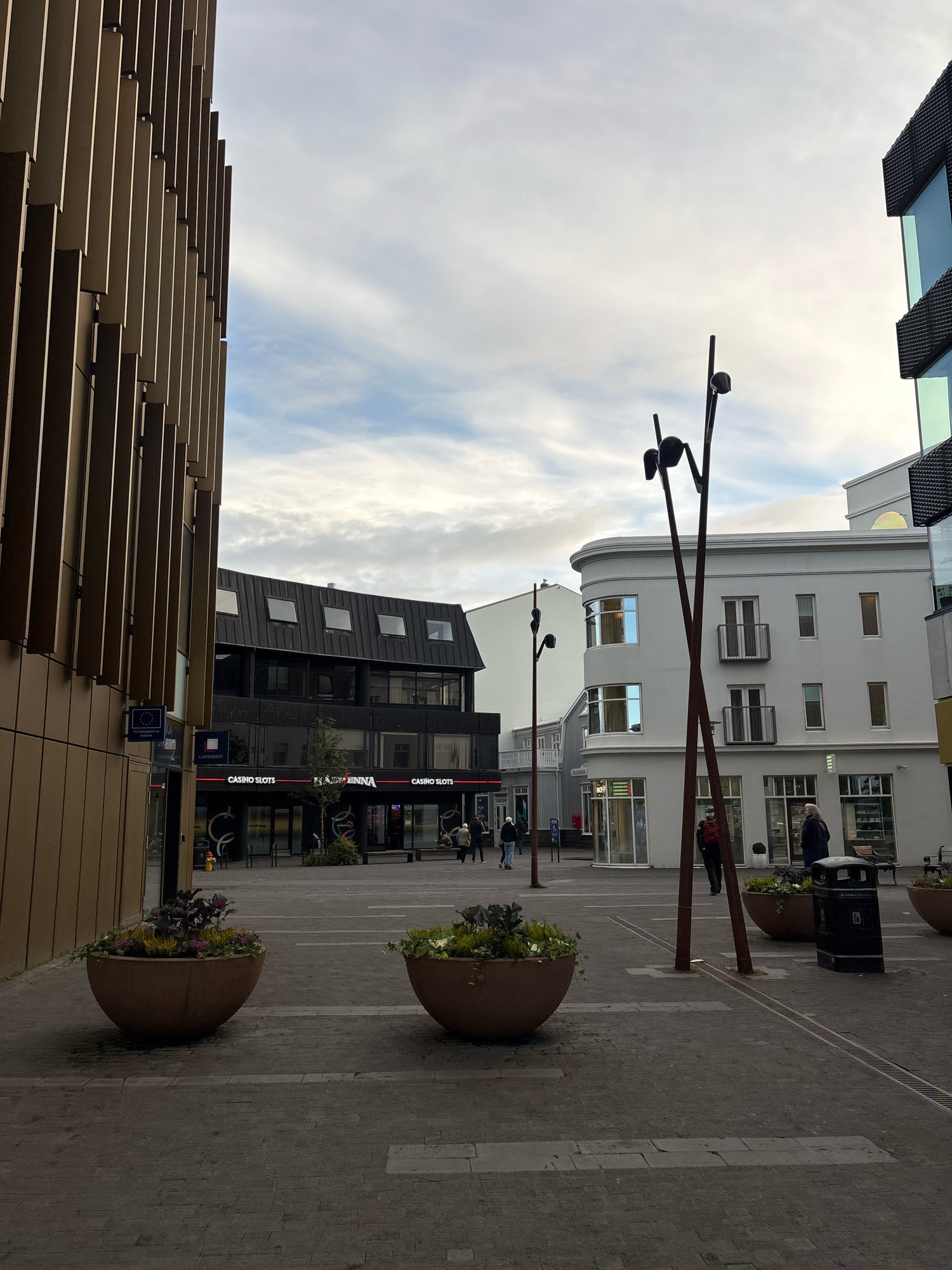 The image depicts a street scene in Reykjavik, Iceland, specifically on Laugavegur street. In the foreground, there are two large, round planters filled with foliage. The ground is paved with cobblestones and includes a metal grate. To the left, a modern building with vertical wooden panels dominates the frame. A blue sign with EU symbols and text is visible on its facade. Further into the scene, a darker, multi-story building with numerous windows and a dark, textured facade is present, featuring signs for "CASINO SLOTS" and "LAGUNA". To the right, a white, rounded building with multiple windows and balconies stands. A tall, abstract metal sculpture with two bulbous elements at the top is positioned in the mid-ground. A black trash receptacle is located near the planters and the sculpture. Several people are walking in the background, some near the buildings and others in the distance. The sky is overcast with clouds.