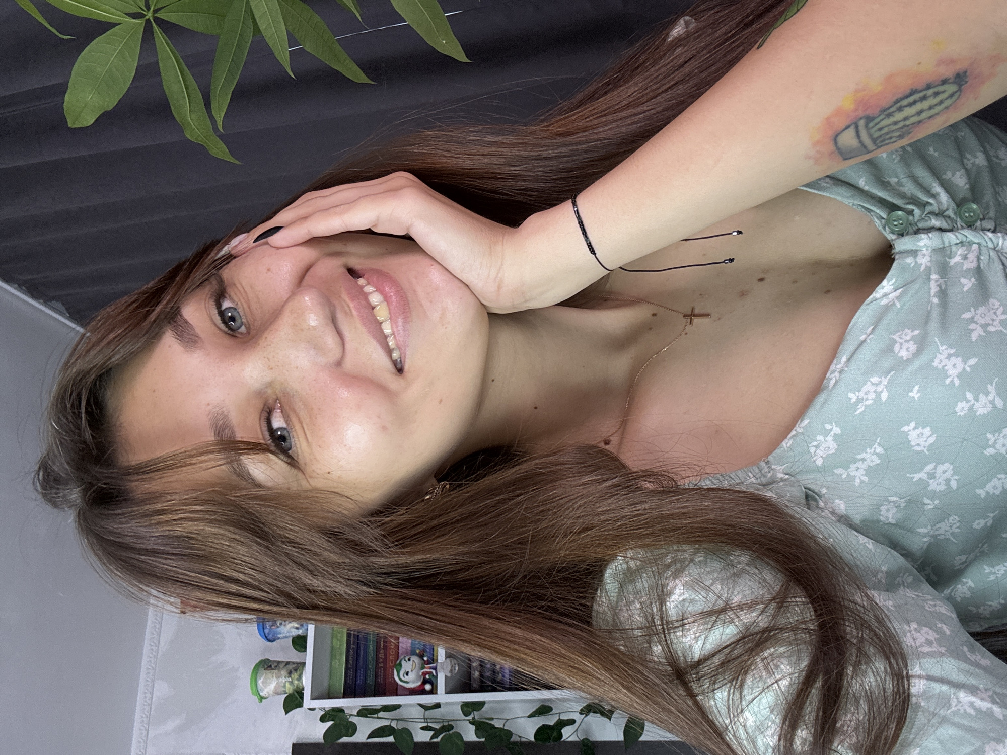 A young woman with long, brown hair and blue eyes is posing for a selfie. She is wearing a light green, floral-patterned dress. She has freckles on her face and her hand is resting on her cheek. On her left arm, she has a tattoo of a jellyfish. She is also wearing a necklace with a cross pendant and a thin black bracelet. In the background, there is a bookshelf with books and decorative items, and a large green plant. The scene appears to be indoors, in a residential setting. There is no information about the specific location within Novokuznetsk, Russia, or any context about the woman's actions or interactions.